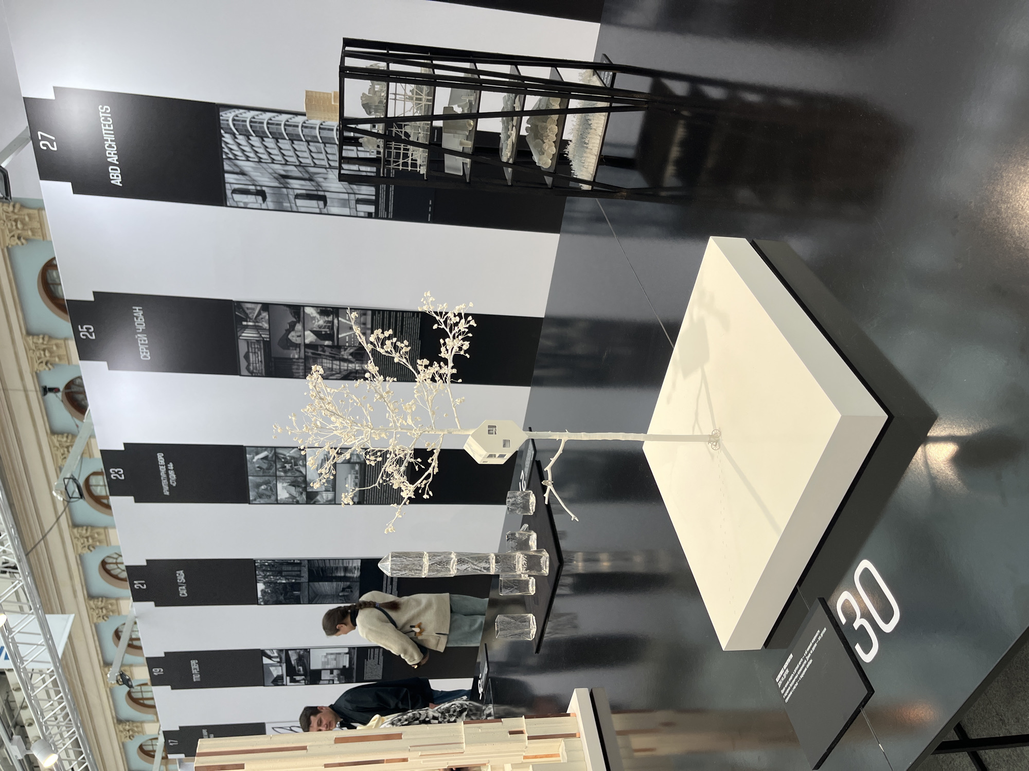 The image shows an architectural exhibition, potentially located in the city of Moscow, Russia, as suggested by text on the exhibits. The exhibition features a series of architectural models displayed on a black surface with numbered white labels, including 17, 19, 21, 23, 25, 27, and 30. The models range from representations of buildings and structures to conceptual art pieces, including a tree with a small house attached to it.
Several people are present, observed near exhibit numbers 17, 19, and 21. The exhibit near number 27 belongs to "ABD Architects" and another project at exhibit number 25 is by "Сергей Чобан" (Sergey Choban).
The environment appears to be well-lit and modern, with elements of the building's architecture, such as arched windows and ornamental details, visible in the background. The lighting fixtures and structure are visible near the ceiling.