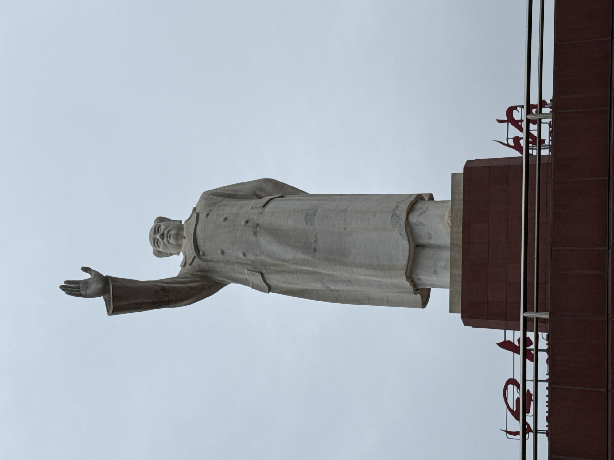 A low-angle, full-shot image captures a tall, light-grey stone statue of a male figure, presumed to be Mao Zedong, against an overcast, light-grey sky. The statue depicts the figure wearing a long, double-breasted overcoat with visible buttons and pockets. His left arm is raised high, palm open, in a gesturing or waving pose. His right arm is bent and positioned behind his back, partially obscured. The figure's head is tilted slightly upward and to the viewer's right. The statue rests on a multi-tiered pedestal. The upper portion of the pedestal is light-grey, matching the statue, while the lower, larger section is composed of reddish-brown rectangular blocks, possibly brick or stone cladding. Below the pedestal, a building or structure with matching reddish-brown facade material is visible. A metal railing, consisting of horizontal bars, extends across the top of this lower structure. Behind the railing, large, stylized red Chinese characters are partially visible, affixed to the top of the reddish-brown structure. The scene is set in Qingyang District, China.
