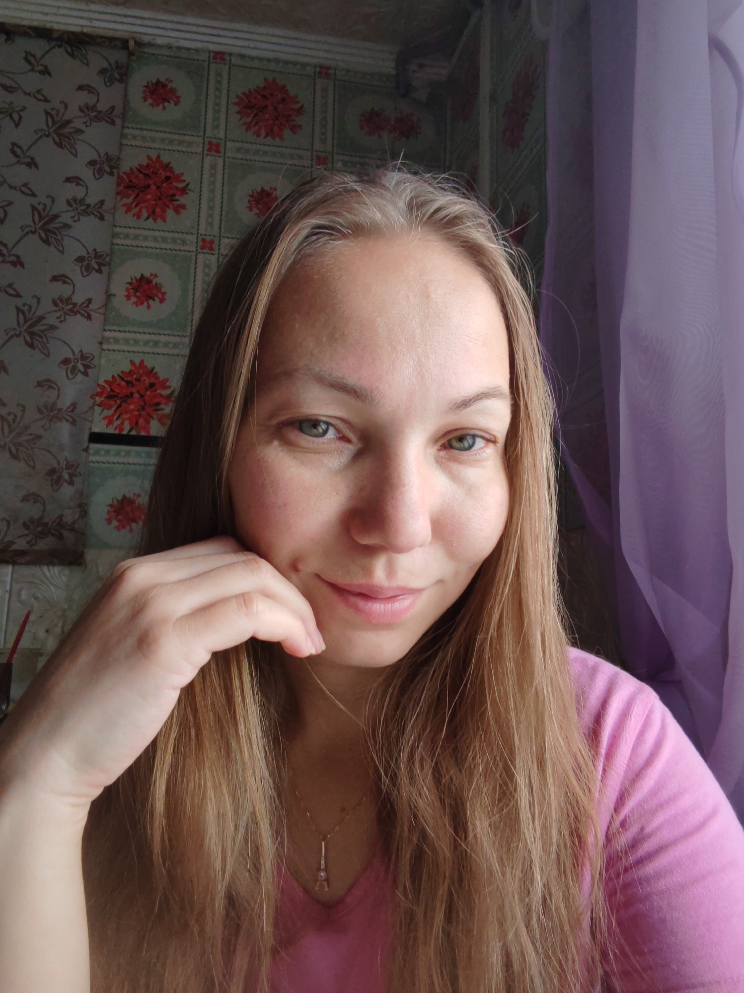 A medium shot shows a woman with shoulder-length blonde hair, parted down the middle, and blue eyes. Her left hand is raised to her chin, with her fingers delicately resting on her cheek. She is wearing a pink v-neck t-shirt. The background features floral wallpaper with red flowers on a green and white grid pattern. To the right, a sheer purple curtain is visible. The lighting suggests an indoor setting near a window, casting a soft glow on her face. The image does not provide any specific location details within the city of Чухлэм, Russia, beyond being an indoor scene.