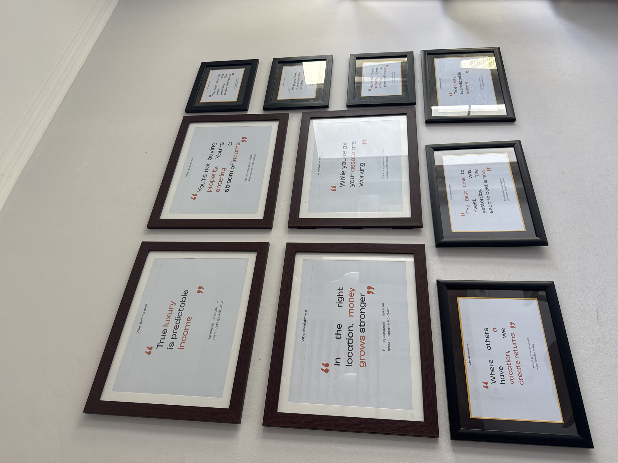 The image shows a white wall with nine framed motivational quotes related to real estate investment, likely displayed in an office or sales environment. The quotes are from "Vibe Development" and are in both English and what appears to be Russian or a similar language. The quotes touch on themes like predictable income, the value of location, and creating returns. The frames are in two colors: dark brown and black. The arrangement is a grid-like formation. There is no information available regarding the city of Denpasar, Indonesia.