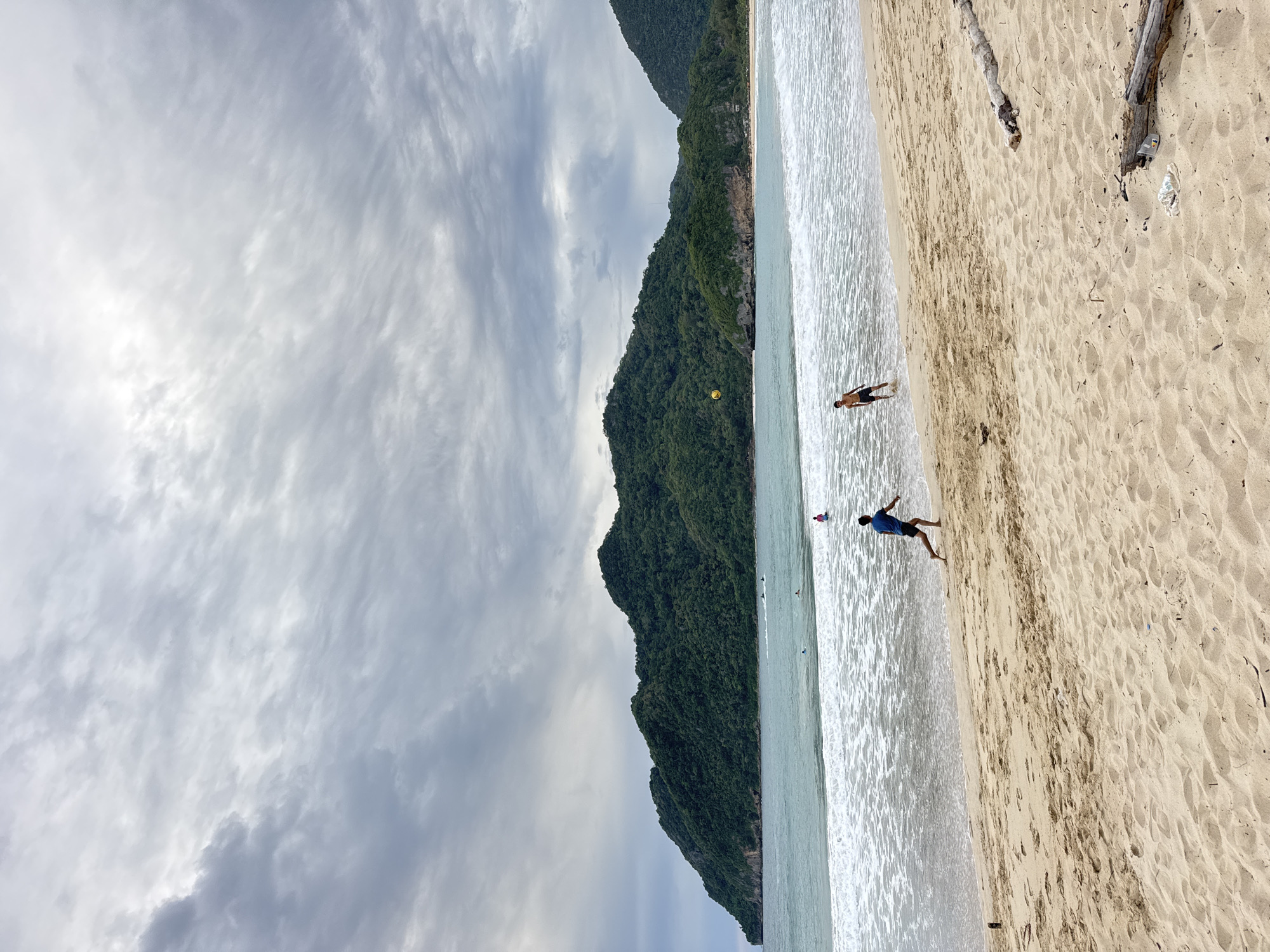 A wide-angle, full shot captures a sandy beach in Meunasahbale, Indonesia, under a cloudy sky. The foreground is dominated by textured sand, with two pieces of driftwood visible on the right. In the middle ground, the ocean meets the shore with foamy waves. Two young men are playing in the shallow water. One, wearing black shorts, runs along the water's edge. The other, in dark shorts, stands further out in the water. A yellow ball is suspended in the air between them. In the background, lush green mountains rise from the sea, with the coastline curving to the right. Several small boats are visible on the water near the shore, and more people are in the water further out. The sky is filled with dramatic, wispy grey clouds.