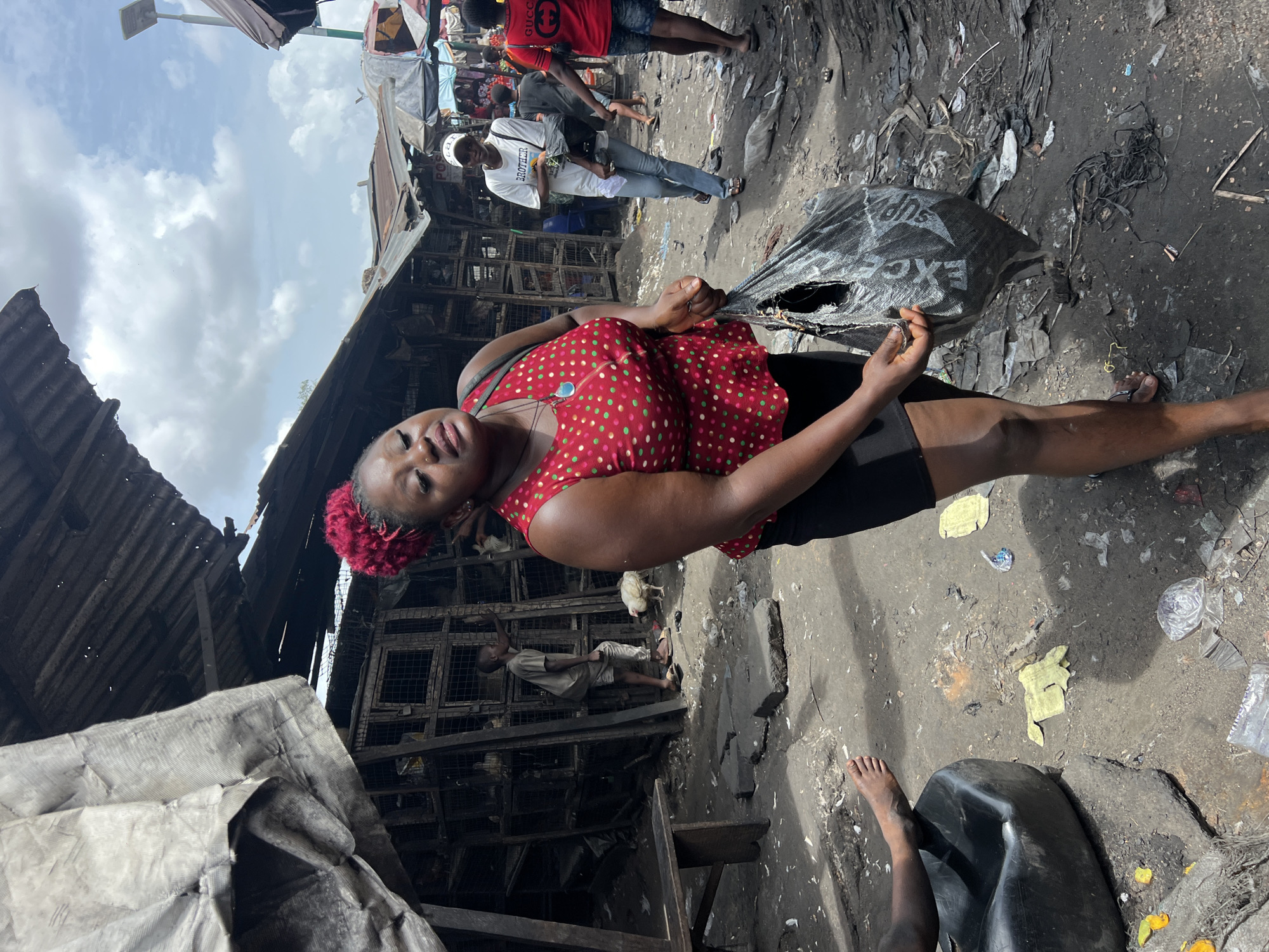A medium, eye-level shot depicts a woman standing in a market in Nkpor, Nigeria. The woman is fair-skinned, wearing a red polka dot top, black shorts, and a red floral headpiece. She is holding a large, grey, torn plastic bag.
The market setting is visibly dirty with debris scattered on the ground. There are indications of damaged stalls, revealing charred wooden structures. Behind the woman, there is a partially visible figure of a person sitting near what appear to be chicken coops, with a single chicken visible. People can be seen in the background, some walking through the market. The sky is partly cloudy, suggesting it is daytime. A black plastic container and a bare foot are visible at the bottom right.