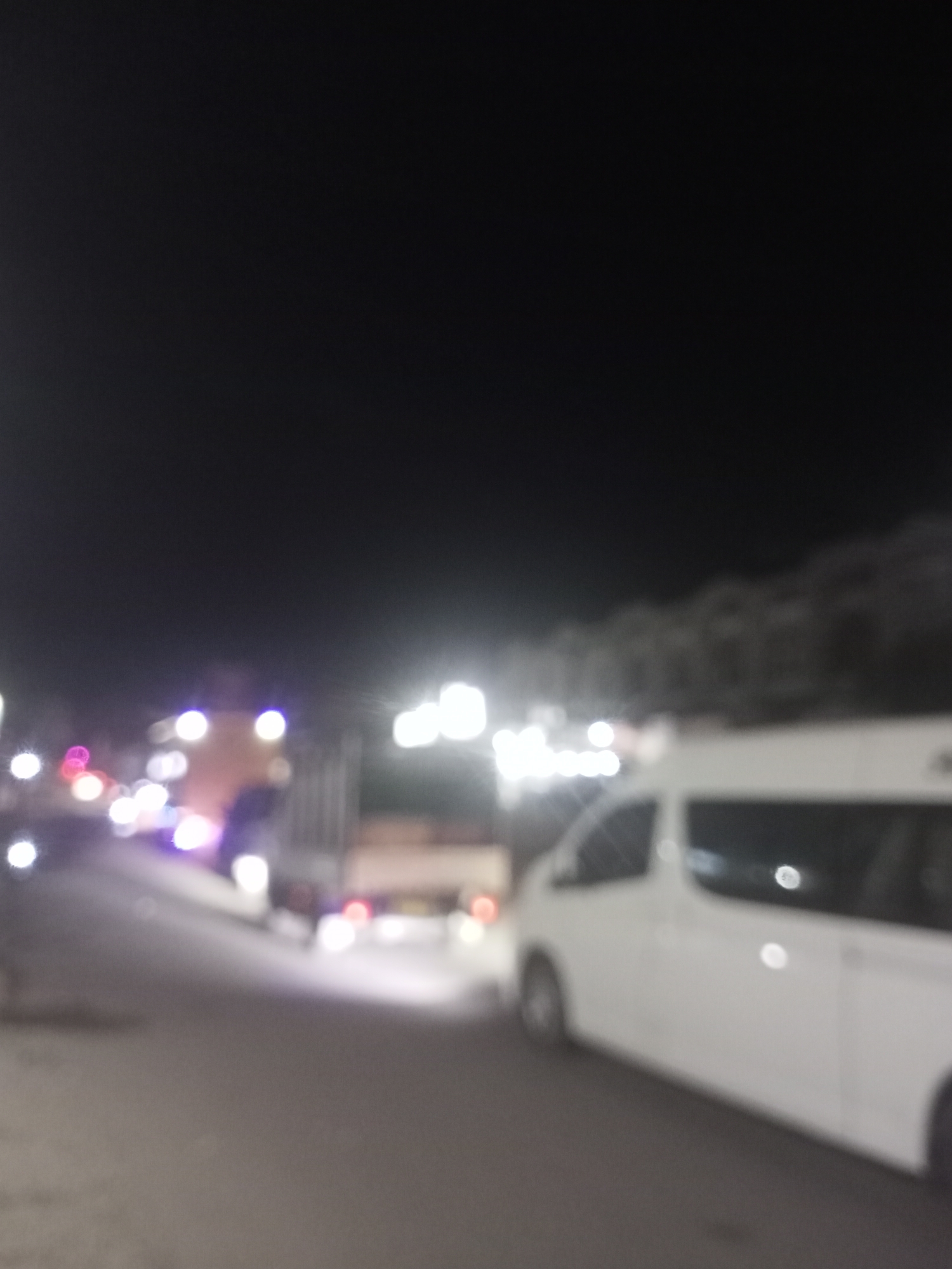 The image is a blurry, low-light photograph taken at night, likely in an outdoor urban setting. The scene appears to be a street with several vehicles present. A prominent white van is visible on the right side of the frame, positioned on a road. Further down the road, a larger truck or bus is partially visible. There are also various lights, some of which appear to be vehicle headlights or streetlights, scattered throughout the scene, contributing to the blurry effect. The background is dark, suggesting it's nighttime, with some indistinct shapes that could be trees or buildings. The overall impression is one of a street scene at night with traffic. The location in Tingkeummanyang, Indonesia, is not visually discernible from the image itself.