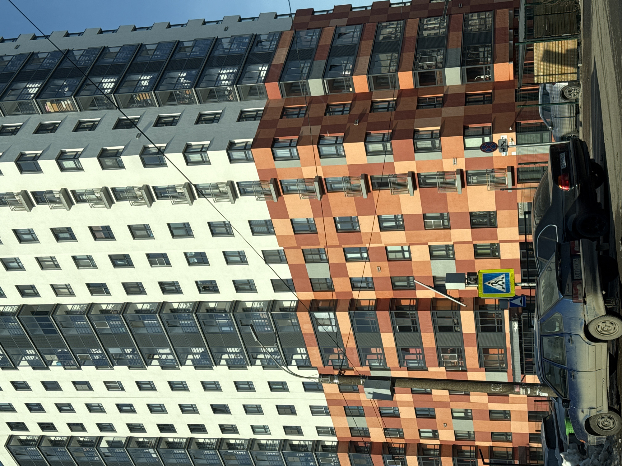 A multi-story building with two distinct facade designs occupies the majority of the frame. The left side of the building features a white facade with rows of windows and balconies, while the right side showcases a checkered pattern of orange and brown tones. A street runs along the right side of the building, where several vehicles, including a blue car and a dark-colored car, are parked. A pedestrian crossing sign is visible on a pole near the cars. The image is taken in Izhevsk, Russia.