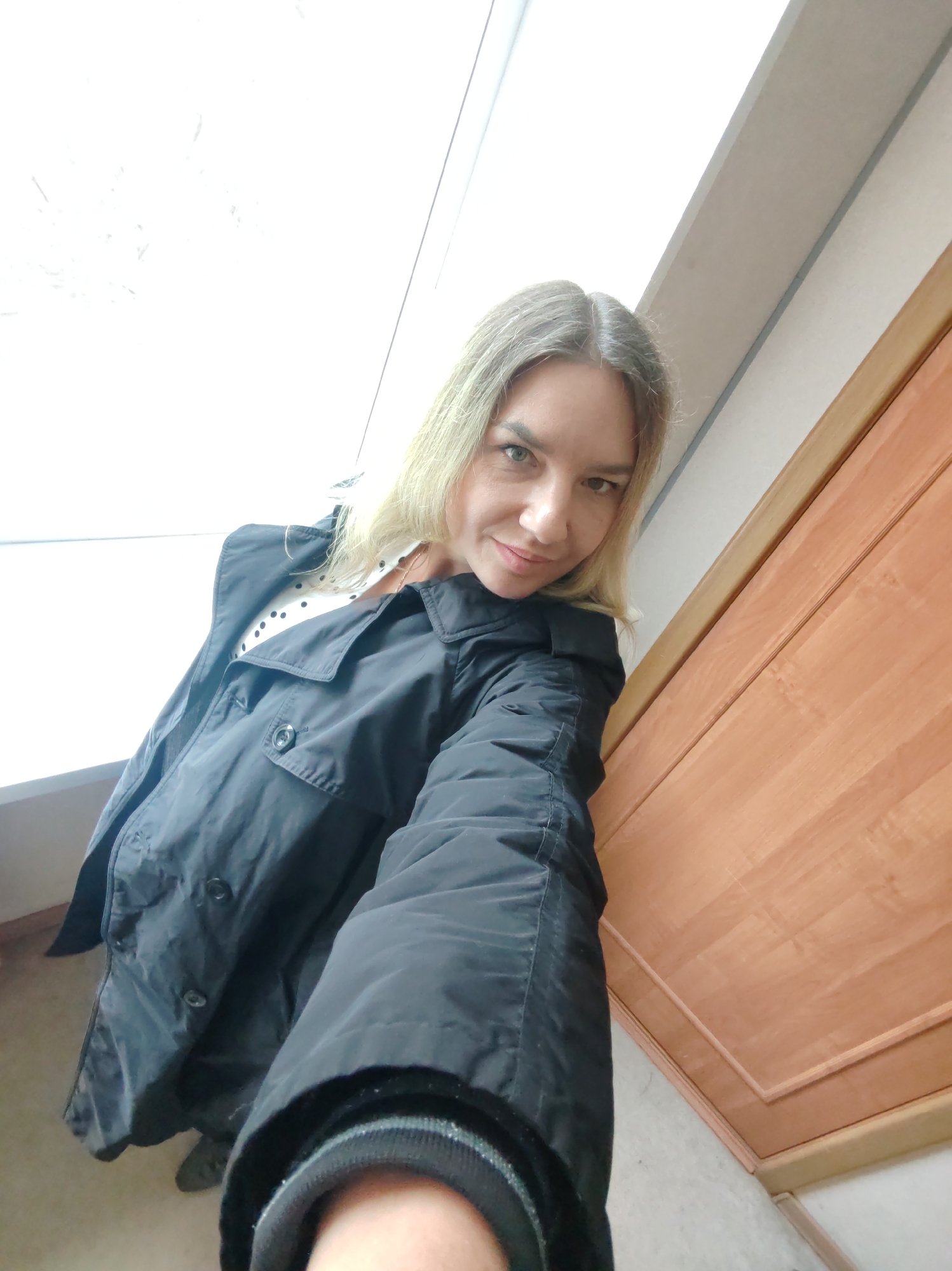 A selfie-style portrait captures a woman from a high angle, looking directly into the camera with a slight smile. She has blonde or light brown hair with a visible part and green/hazel eyes. She is wearing a black collared jacket or coat over a white shirt with black polka dots, which is visible at her chest. Her arm, holding the camera, is extended towards the bottom right of the frame, showing the cuff of her jacket.
The background consists of a bright, overexposed white area on the left, likely a window or a brightly lit wall, and a light brown wooden door with raised paneling on the right. A light-colored floor is visible at the bottom of the image. The lighting suggests it is daytime, with bright light coming from the left. There is no visible text in the image. The setting appears to be indoors, possibly a hallway or room.
