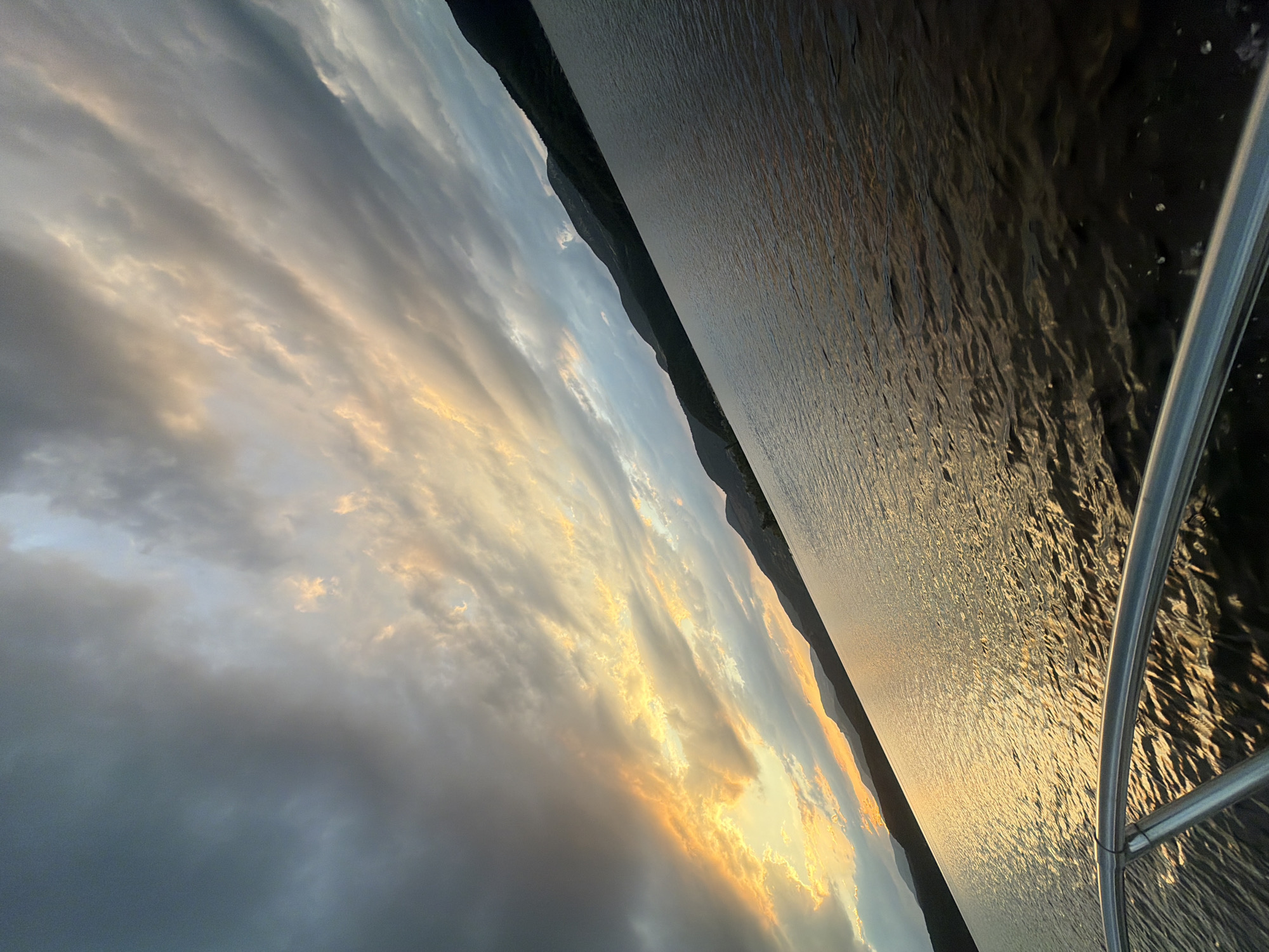 The image depicts a scenic view of the waters of the Glavatartsi dam in Glavatartsi, Bulgaria, during what appears to be sunset or sunrise. The sky is filled with dramatic clouds, with a band of bright yellow and orange light breaking through from the left, indicating the sun's position. The water reflects the colors of the sky, creating a shimmering effect. Dark, silhouetted hills form the background along the horizon. In the foreground, a silver railing suggests the image was taken from a boat or a pier. There are no people visible in the image. The overall mood is tranquil and majestic, capturing the natural beauty of the location.