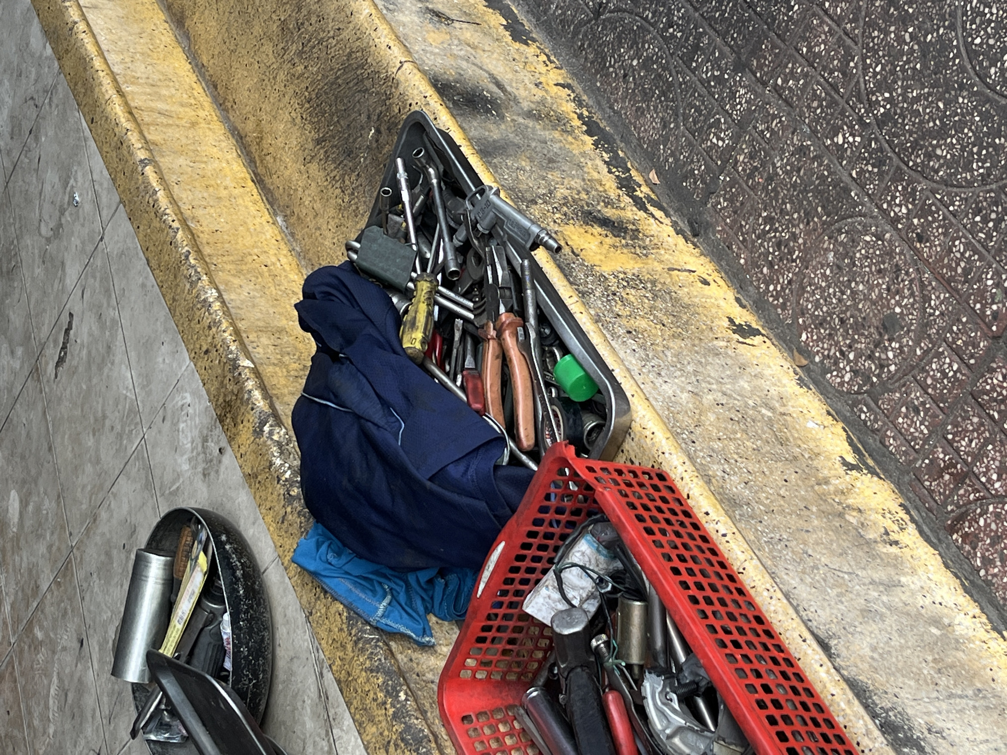 On the edge of a sidewalk in Bac Nha Trang Ward, Vietnam, a collection of tools and equipment rests in disarray. A metal tray, partially covered by a folded blue garment, holds an assortment of tools including wrenches, screwdrivers, and pliers. Adjacent to the tray, a red plastic crate contains additional items such as metal cylinders, wires, and other unidentified mechanical components. To the left, a motorcycle part, possibly a fender or side panel, suggests the tools might be for motorcycle repair. The sidewalk is bordered by a yellow curb and a section of patterned paving stones.