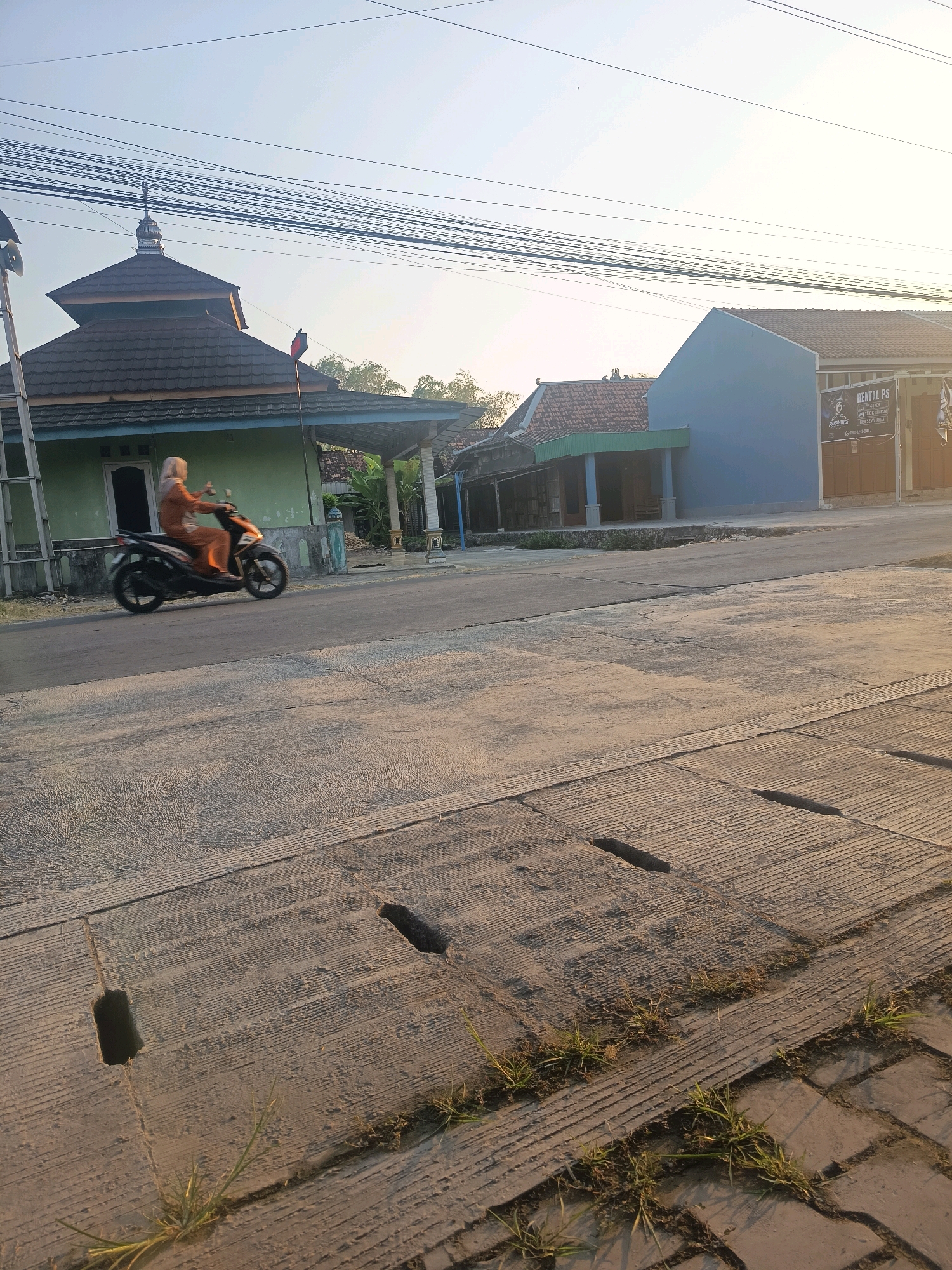 In Kalisari, Indonesia, a woman wearing an orange headscarf and an orange outfit rides a black and orange scooter on a paved road. The scooter is blurred due to motion. To the left of the road, there is a green building with a brown roof and a dome, possibly a mosque, with a flagpole in front. Further down the road, there are more buildings, including a blue building with a sign that reads "RENTAL PS". The road surface in the foreground is concrete with visible drainage grates. The scene appears to be in a residential or commercial area during daytime, with utility wires overhead.