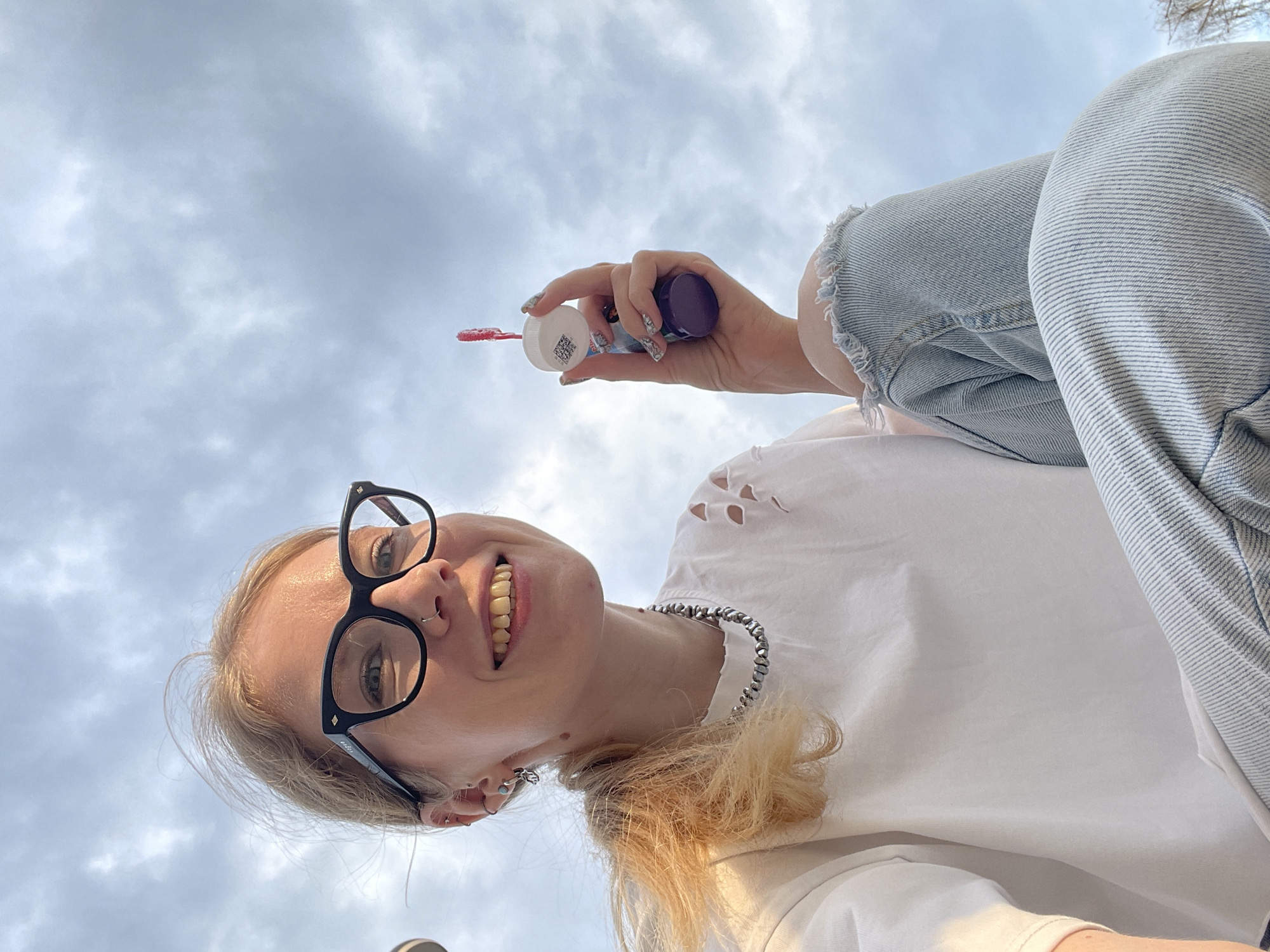 The image depicts a young woman with blonde hair, wearing black-framed glasses and a white t-shirt with distressed details. She has a nose ring and multiple earrings. She's holding a bottle of bubble solution with a red wand. She is wearing ripped jeans. The background consists of a cloudy sky. There is no evidence to suggest this photo was taken in Izhevsk, Russia.