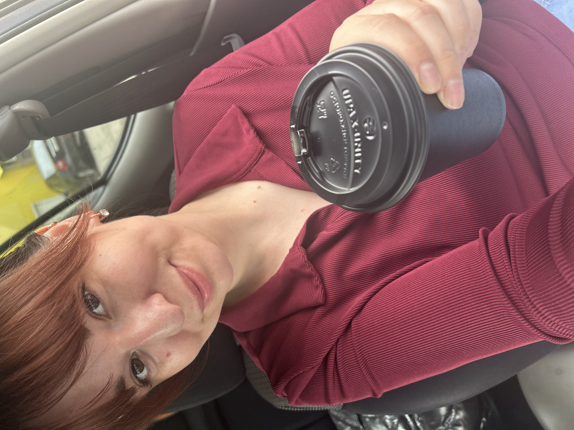 A woman with fair skin, auburn hair with bangs, and brown eyes is depicted in a selfie, positioned within the interior of a vehicle. She wears a deep maroon, ribbed, collared, long-sleeved shirt and small, clear stud earrings. A faint smile is present on her lips. She extends her right hand, holding a black disposable coffee cup with a black plastic lid, towards the camera. The lid displays reversed text, including "UPA-X-UNITY" and "1.25". The car interior features dark upholstery, visible on the headrest behind her, and a grey seatbelt assembly to the right. Through the car window, an exterior scene is visible: a section of a bright yellow building or wall is present on the left, and a dark-colored sedan is parked further down a street. The overall environment suggests an urban setting, specifically in the city of Chaikovsky, Russia.