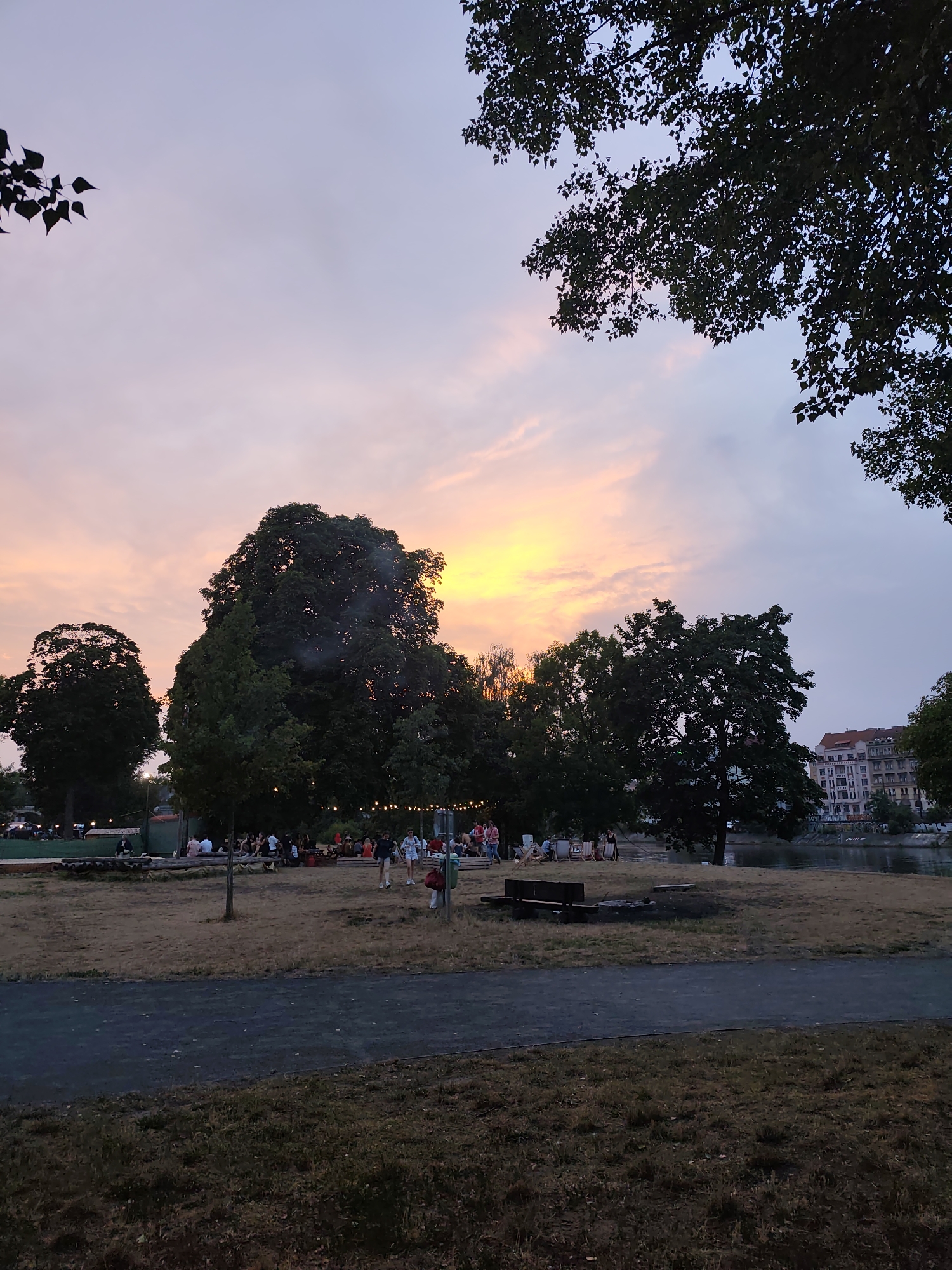 The image depicts a sunset scene in Prague, Czechia, likely at a riverside park or beer garden. The sky is a gradient of soft pinks and oranges, transitioning to a pale blue, with the sun setting behind silhouetted trees and buildings. In the foreground, a grassy area is bordered by a dark asphalt path. Further back, a group of people are gathered on a sandy or gravelly area, some sitting on logs and chairs, others standing and conversing. There are several trees, some large and leafy, scattered throughout the scene. In the distance, buildings line a body of water, likely the Vltava River, with a waterfront promenade visible. A bench and what appears to be a fire pit are also present in the mid-ground. The overall atmosphere is relaxed and social, with people enjoying the evening outdoors.