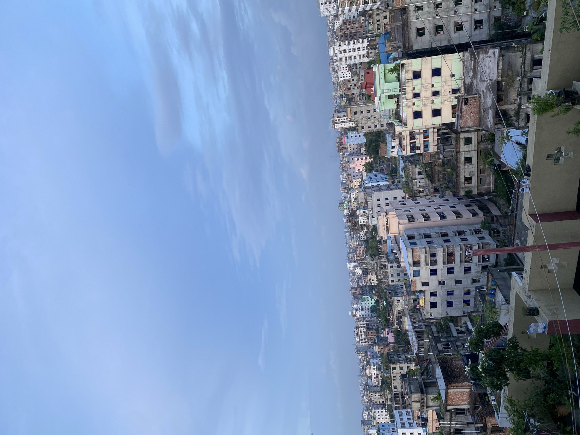 The image depicts a panoramic view of a densely populated urban area in Dhaka, Bangladesh, likely a residential district. The perspective is from a high vantage point, looking over a multitude of multi-story buildings. The architecture is varied, with many structures appearing to be in different stages of construction or in a state of moderate disrepair. Buildings are predominantly concrete, with a mix of pastel and neutral colored facades, some showing exposed brickwork. Balconies and rooftop structures are visible on many buildings, with some rooftops appearing to be used for vegetation or storage. The sky above is a muted blue with scattered, wispy clouds. In the foreground, a rooftop area is visible with some potted plants, a water tank, and some personal belongings like shoes. The overall impression is of a bustling, active cityscape characterized by dense housing and a developing urban environment.