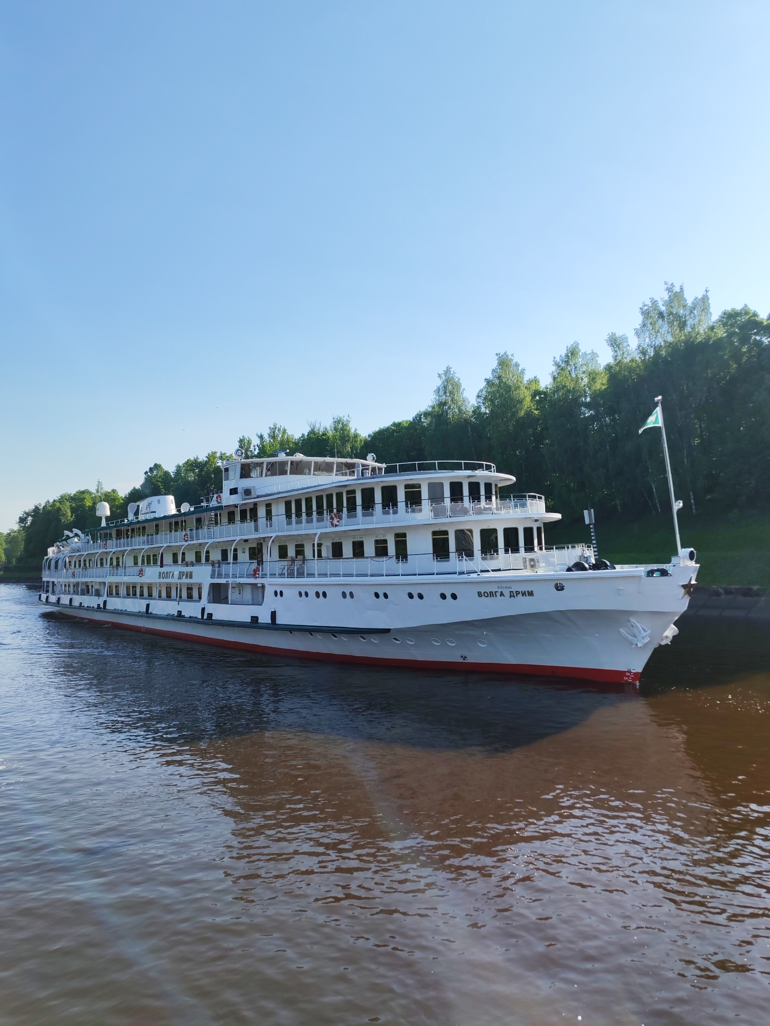 The image depicts the Volga Dream, a white river cruise ship, on a body of water, likely a canal or river, in Iksha, Russia. The ship is white with multiple decks and the words "VOLGA DREAM" written on its side. A green flag is mounted at the front of the ship. The water is brown and reflects the ship and the sky. The background shows a green landscape with trees lining the waterway. The sky is clear blue.
