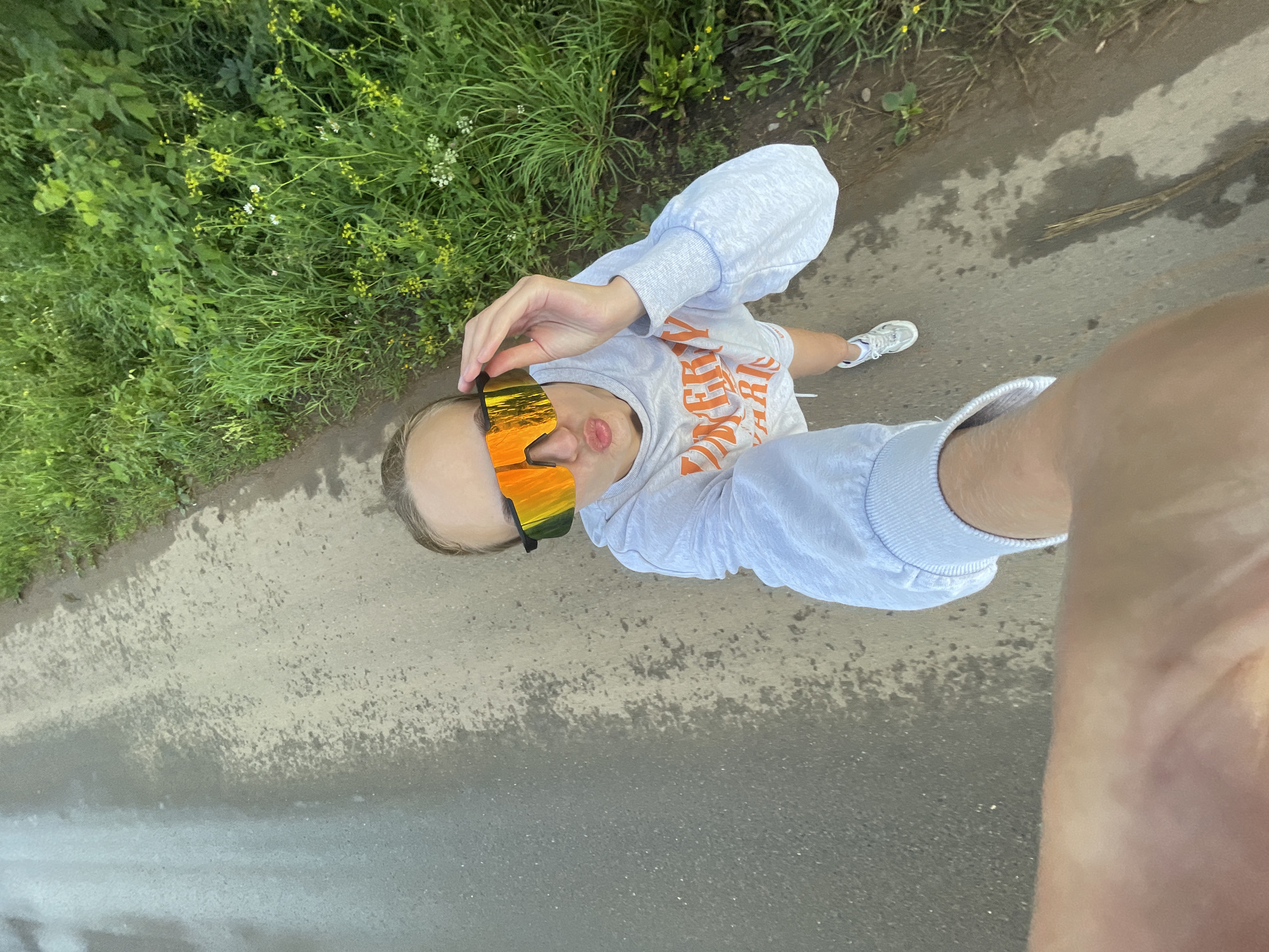 A first-person perspective shot shows a person wearing reflective sunglasses with orange and yellow lenses, a light grey sweatshirt with orange text, and white athletic shoes. The person is standing on a wet asphalt surface next to a grassy area with small yellow and white wildflowers. The scene appears to be outdoors. The image does not provide any specific information about the location within the city of Подшивалово, Russia.