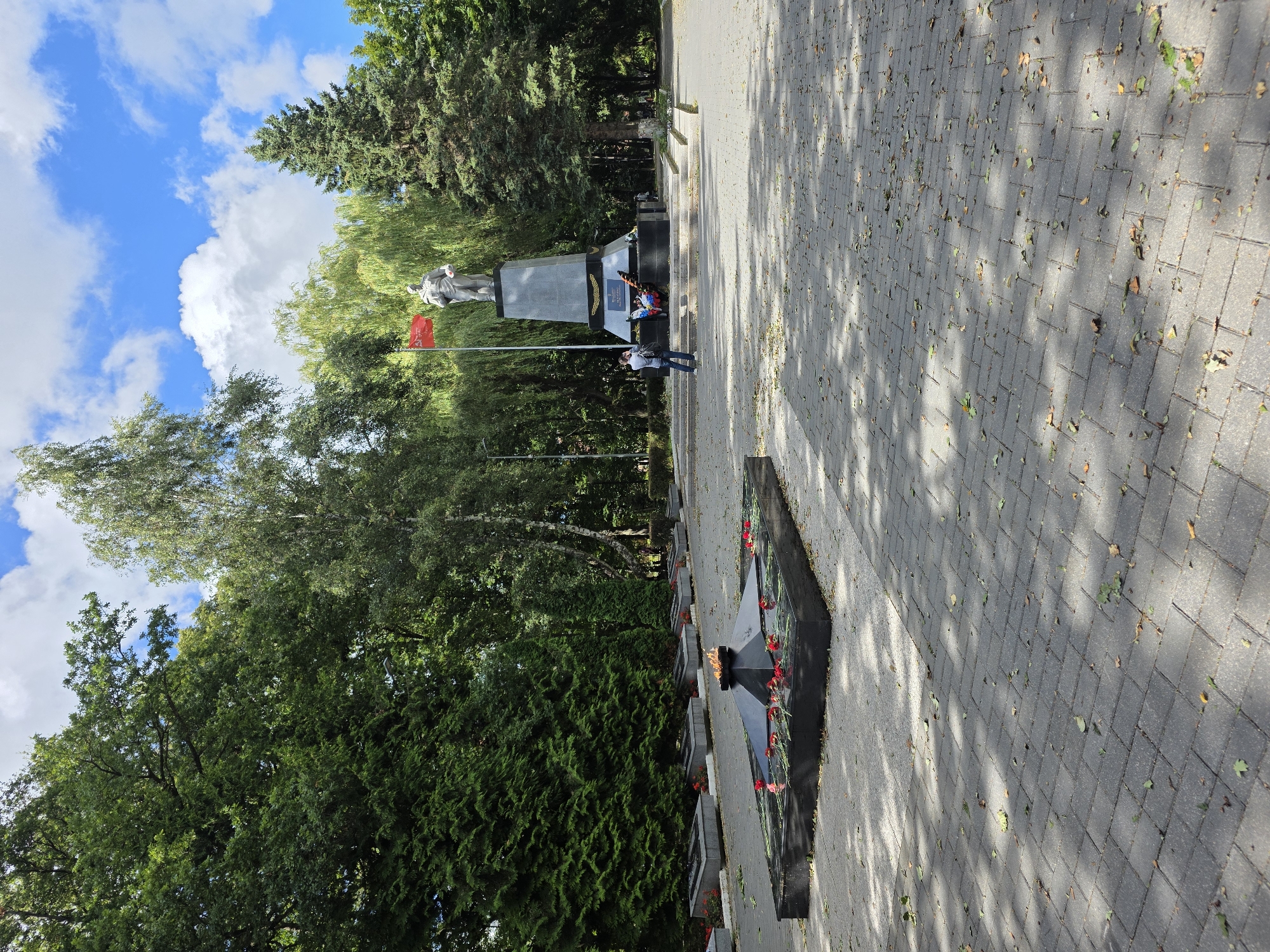 The image depicts a memorial complex. In the foreground, an eternal flame burns atop a black granite structure, with red flowers laid around its base. To the left, a row of granite grave markers is visible, also adorned with red flowers. In the background, a large statue of a man stands on a pedestal, topped with a red flag. Two people are present: one, a person in a light-colored shirt and dark pants, is standing near the statue, appearing to take a photograph. Another person is visible in the distance. The scene is set within a park or public space with lush green trees and a paved walkway made of interlocking bricks, with scattered leaves on the ground. The sky above is blue with white, fluffy clouds. This memorial is located in Zelenogradsk, Russia.