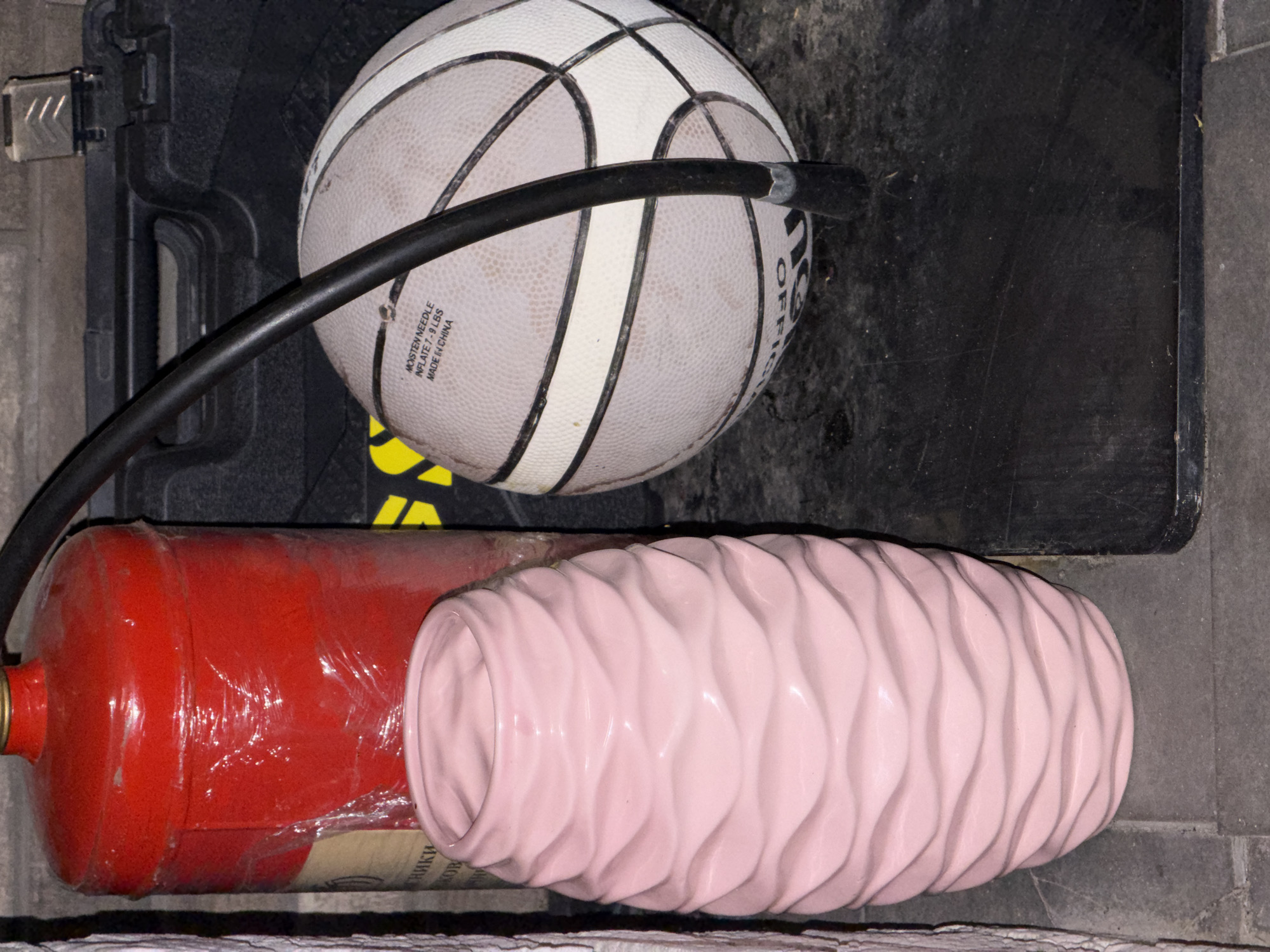 In a dimly lit, possibly storage area, a red fire extinguisher wrapped in plastic is positioned vertically on the left. Adjacent to it, a pale pink, wavy textured vase stands on the floor. To the right, a black container, possibly a case, is visible. Resting on the case is a basketball, partially obscured by a black tube connected to a pump or hose. The floor appears to be tiled. There is no indication of any specific location within the city of Bila Tserkva, Ukraine.