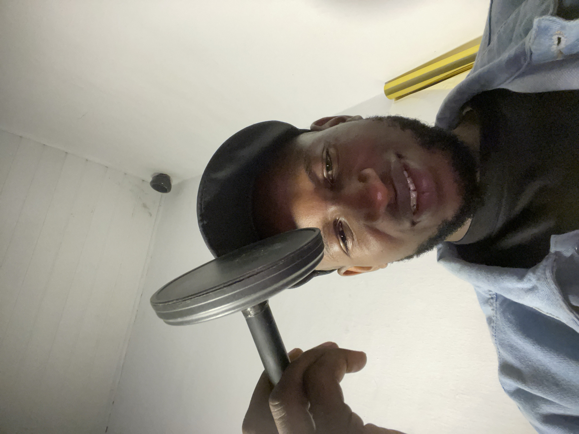 A man wearing a black cap and a denim jacket is holding a dumbbell above his head. He is looking down at the camera with a slight smile. The background is a white wall with some details on the ceiling and a yellow object leaning against the wall. The image appears to have been taken indoors, possibly in a residential setting in Durumi, Nigeria.