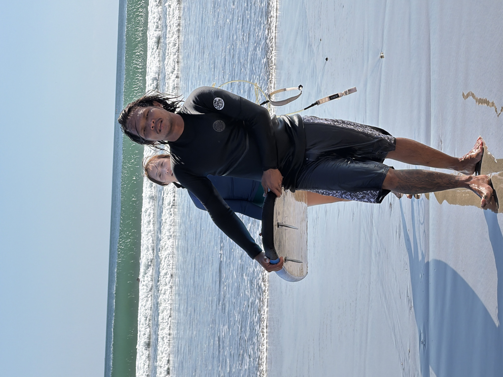 Two individuals, a male and a female, are on the beach in Kuta, Indonesia. The male, in the foreground, is wearing a black long-sleeved rash guard and patterned swim trunks. He is holding a white surfboard. The female, behind him, is wearing a blue rash guard. The ocean is visible with waves breaking in the background. The sky is clear and blue. The ground is wet sand reflecting the light.