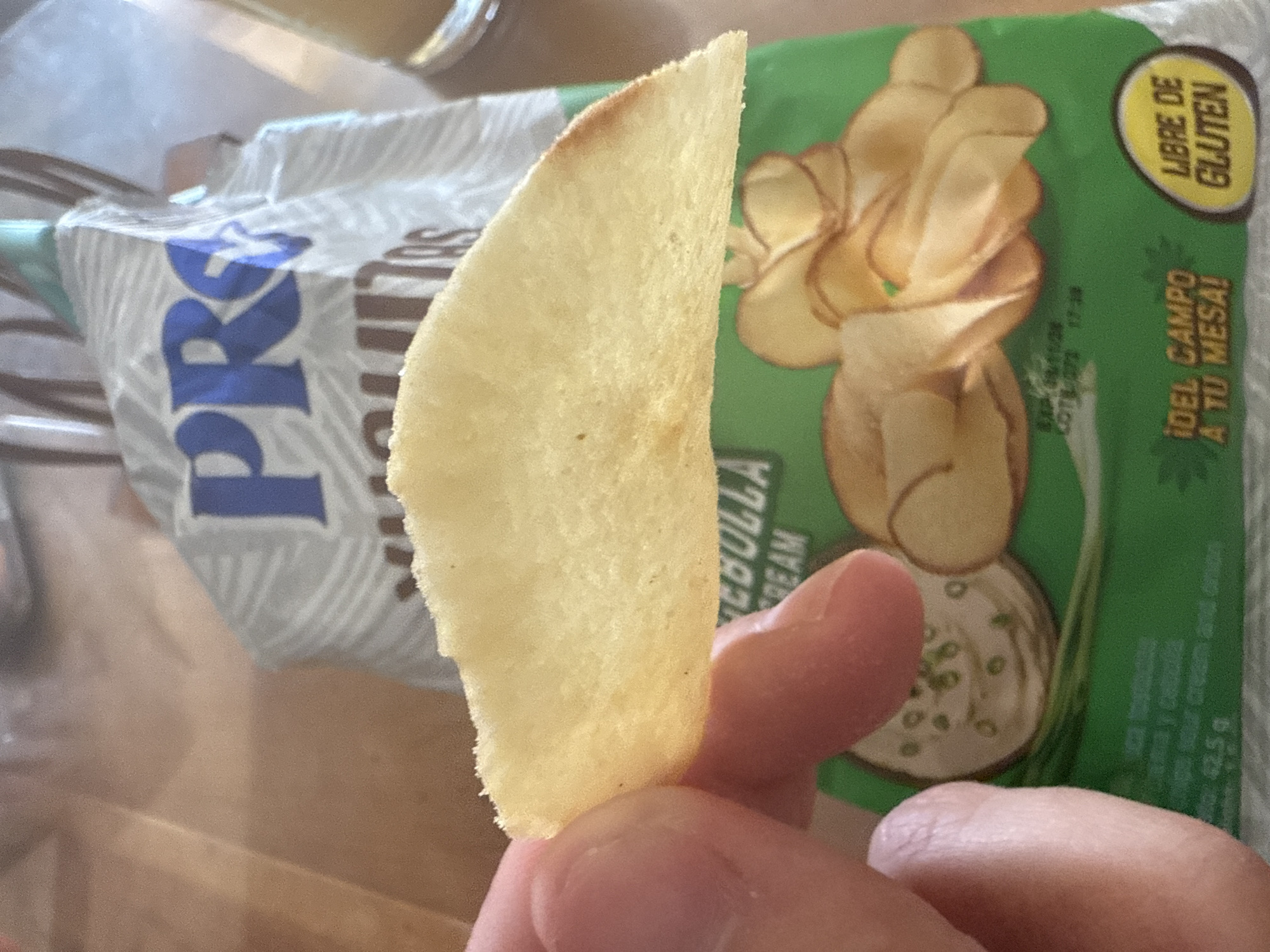 A hand holds a curved potato chip in front of a green package of "PRG" brand potato chips. The chip is yellowish in color, and the package indicates the chips are "Cebolla Cream" flavored. The package also indicates "Libre de Gluten" (gluten-free) and includes the text "¡DEL CAMPO A TU MESA!" The location is unspecified, but the "PRG" brand potato chips and Spanish language on the packaging suggest the image was taken in a location where these products are available, potentially in Xochitepec, Mexico.