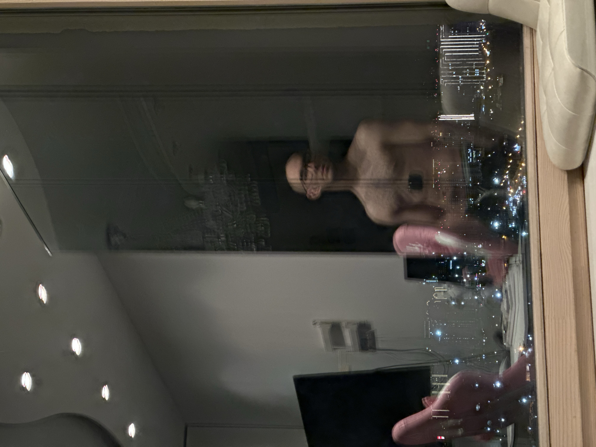 A person stands shirtless near a window. The reflection in the window shows the interior of a room with ceiling lights and a television. Through the window, a cityscape is visible, suggestive of an urban area at night. The location appears to be in Moscow, Russia.