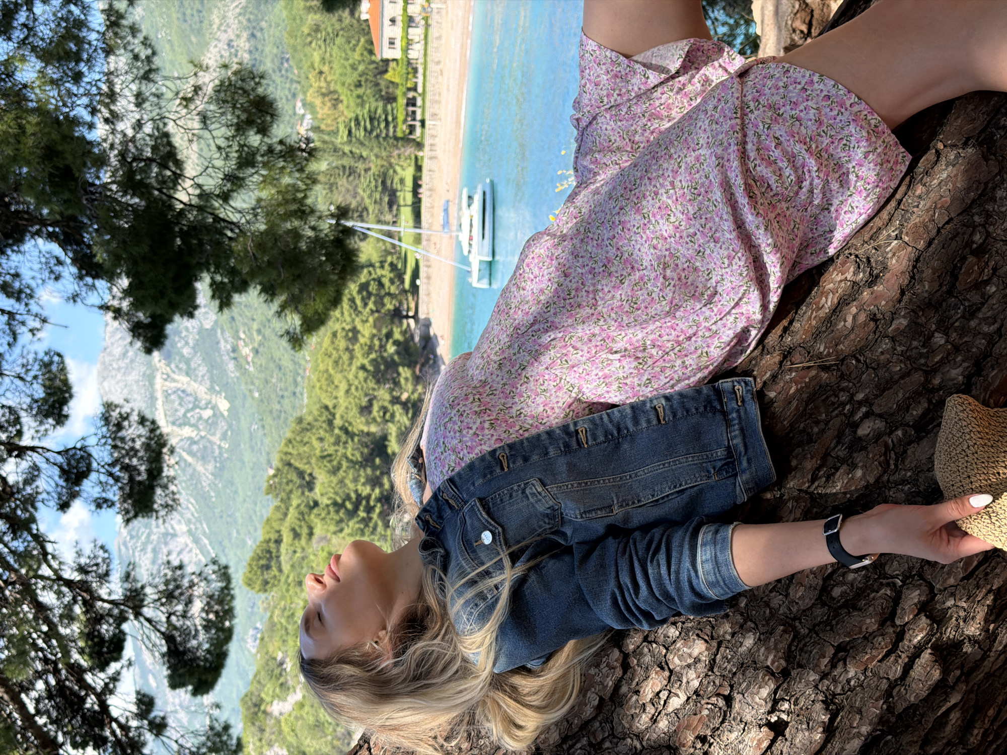 A woman is lying on a tree branch, with a view of Sveti Stefan, Montenegro in the background. She is wearing a floral dress and a denim jacket, and is holding a straw hat in her right hand. She is also wearing a black wristwatch. The background shows the beach, turquoise water, a boat, buildings, trees, and mountains.