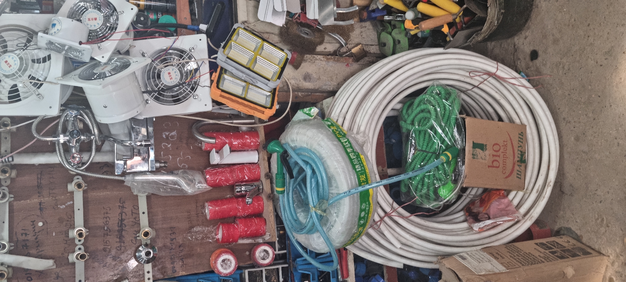 The image depicts a cluttered store stall, likely in Bokhtar, Tajikistan, filled with various plumbing supplies and electronic fans. Prominently displayed are large coils of white plastic tubing and a coiled blue garden hose. Several smaller, red-wrapped cylindrical items, possibly filters or sealants, are arranged on a shelf alongside a white and chrome faucet and a metallic faucet. Above these, a chrome shower fixture with multiple spray settings and a flexible hose is mounted.
A collection of white square electronic fans with visible blades and wiring are stacked and suspended in the upper portion of the image. Two rectangular, orange and gray light fixtures with multiple illuminated panels are also present, hanging from wires.
In the lower right, a woven basket contains an assortment of tools and yellow-handled items. To the left, a cardboard box with visible text and product labels is placed on the floor. A small brown box with "bio komplekt" and Cyrillic text is situated near the coiled tubing.
The overall environment appears to be an indoor market or workshop, with a rough wooden back wall and a concrete floor. The lighting is uneven, suggesting the stall is dimly lit with some artificial light sources. No people are visible in the frame.