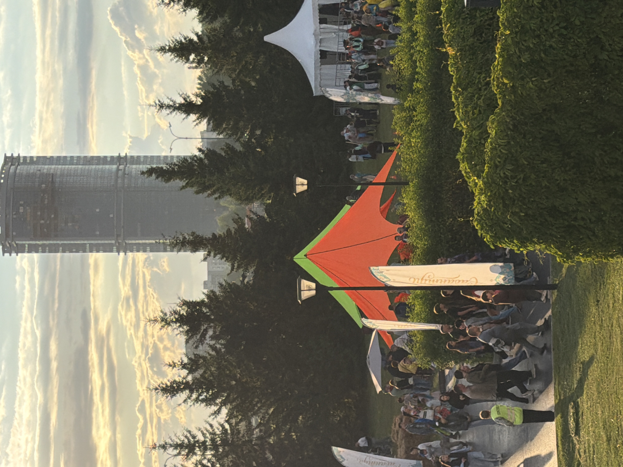 The image depicts an outdoor event, possibly a festival or fair, set against a backdrop of trees and a tall, modern skyscraper under construction. The foreground is dominated by lush green bushes and a grassy area. Several people are visible, some walking, others gathered around tents and stalls.
In the center of the frame, a distinctive orange and green tent structure is set up, with a lamppost in front of it. To the right, a large white tent with an arched roof is visible, with many people milling around it. Flags with text are placed at intervals throughout the scene. The sky is filled with dramatic clouds, illuminated by what appears to be late afternoon or early evening sunlight, casting a warm glow on the scene. The skyscraper in the background, though unfinished, is a prominent feature of the urban landscape.