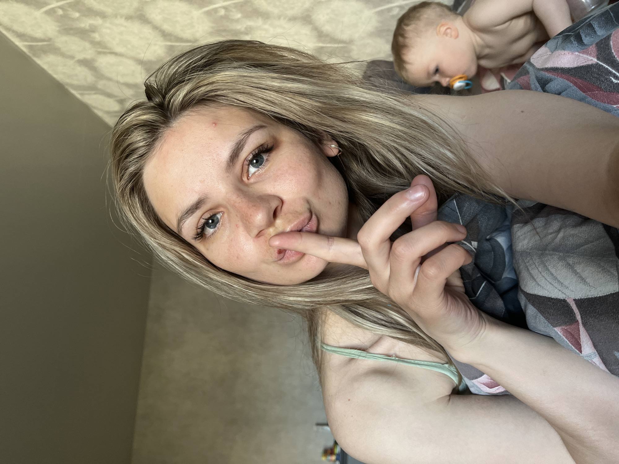 A blonde woman is lying down, holding a finger to her lips in a "shh" gesture. Behind her, on the same bed, is a baby with a pacifier. The location is unspecified, and there is no indication that the image was taken in Chaikovsky, Russia. The background consists of a patterned wall.