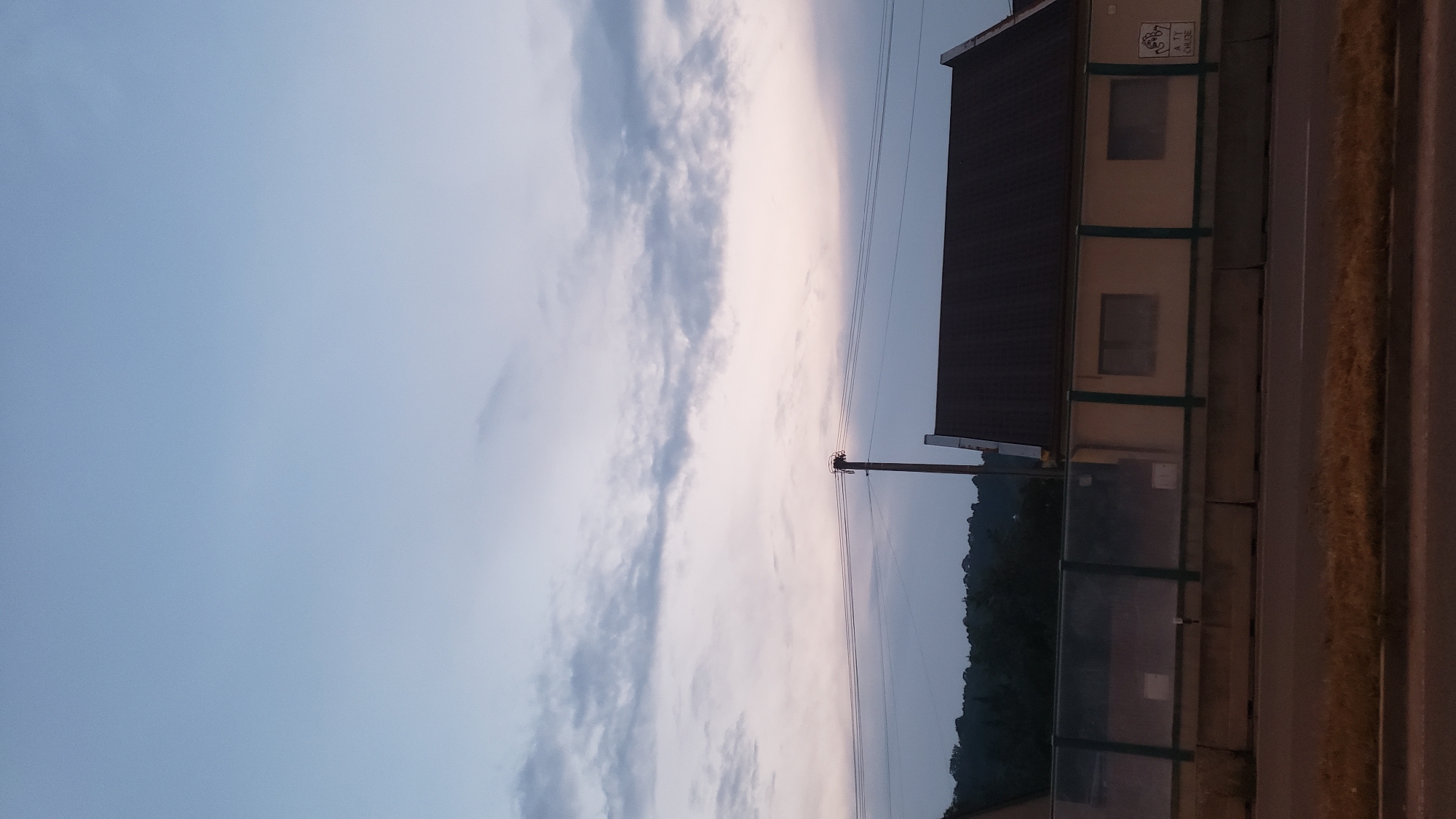A low-angle, vertical shot captures a pale sky with wispy clouds at dusk. Below the sky, power lines stretch horizontally, supported by a single utility pole. In the mid-ground, a low building with a dark, sloped roof is visible, featuring a light-colored facade with two windows. A green-framed transparent barrier runs along the front of the building. Concrete barriers line the foreground, separating the building area from a road with dry grass on its shoulder. The background features a dark, tree-covered hillside. This scene is consistent with the vicinity of the D1 motorway near Prešov, Slovakia, possibly near an overpass or an adjacent service area.