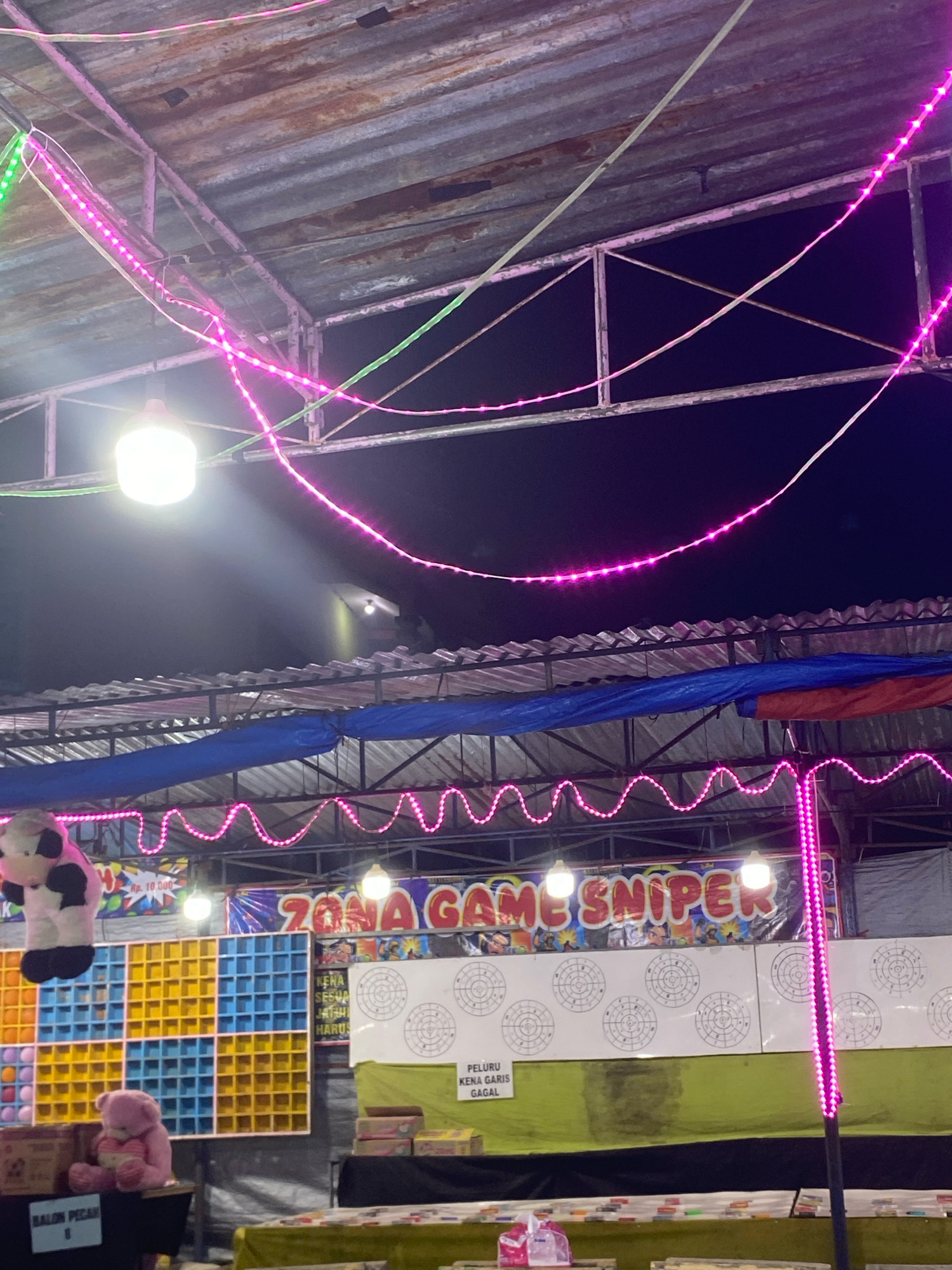 The image depicts a night scene at what appears to be an outdoor fair or carnival in Rejosari, Indonesia. The foreground shows a table with some boxes and a pink plush bear. To the left, a sign labeled "BALON PECAH" is visible. Behind the table is a game stall with a grid of yellow and blue squares, possibly for a prize redemption game, and a large stuffed cow toy hanging above it. Further back, a banner reads "ZONA GAME SNIPER," indicating a shooting game. The background features a white board with several circular targets, and below it, a green tarp. The area is illuminated by various lights, including strings of pink LED lights draped from the corrugated metal roof structure and some hanging light bulbs. The overall environment suggests a recreational setting within the city of Rejosari.