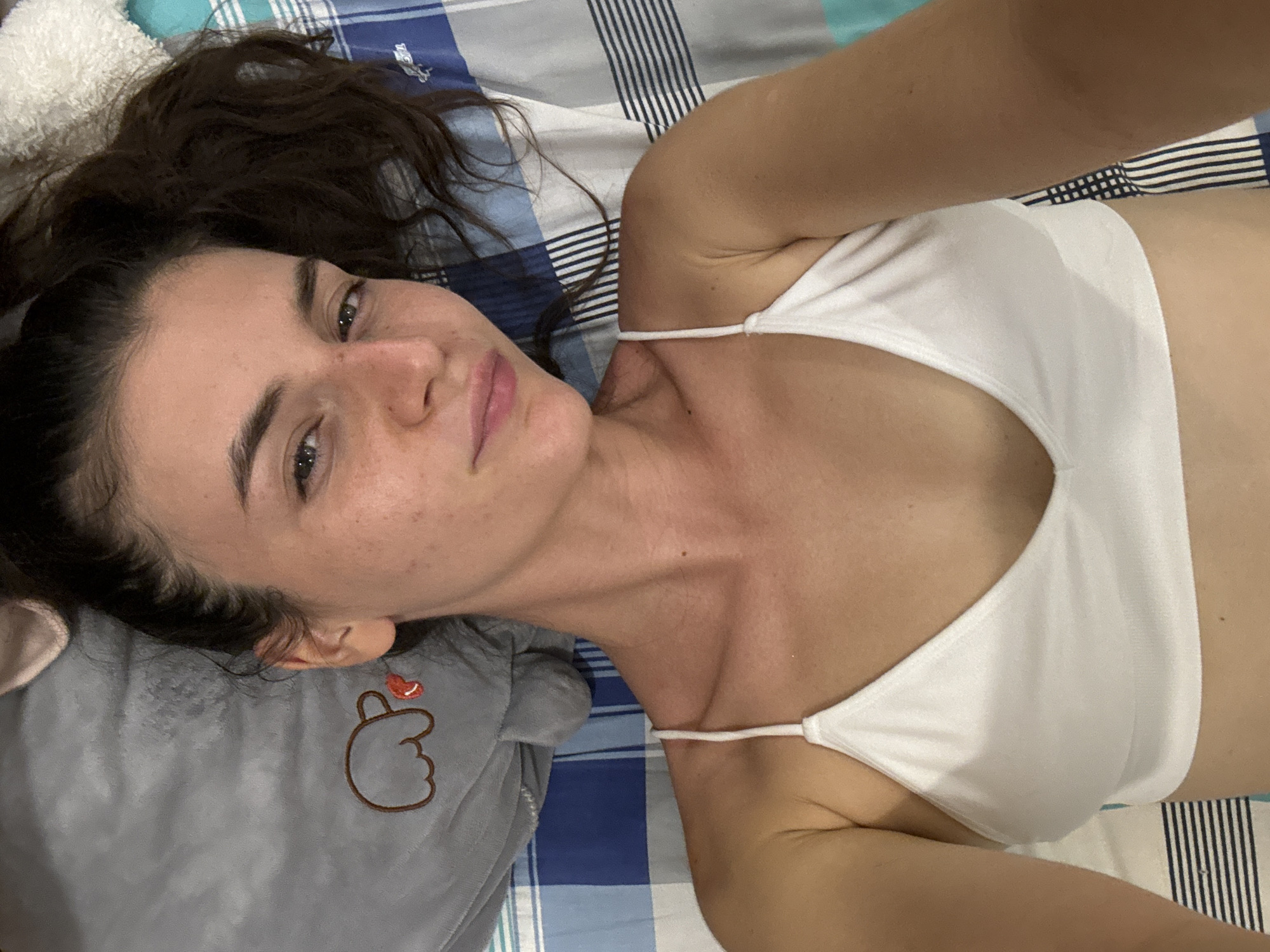 A woman with dark hair and light skin is lying on a bed with a blue and white striped sheet. She is wearing a white top with thin straps. Her head is resting on a gray stuffed animal with a heart-shaped design embroidered on it. The photo appears to be taken indoors, in a room. No specific location in the city of Mũi Né, Vietnam can be determined from the image.