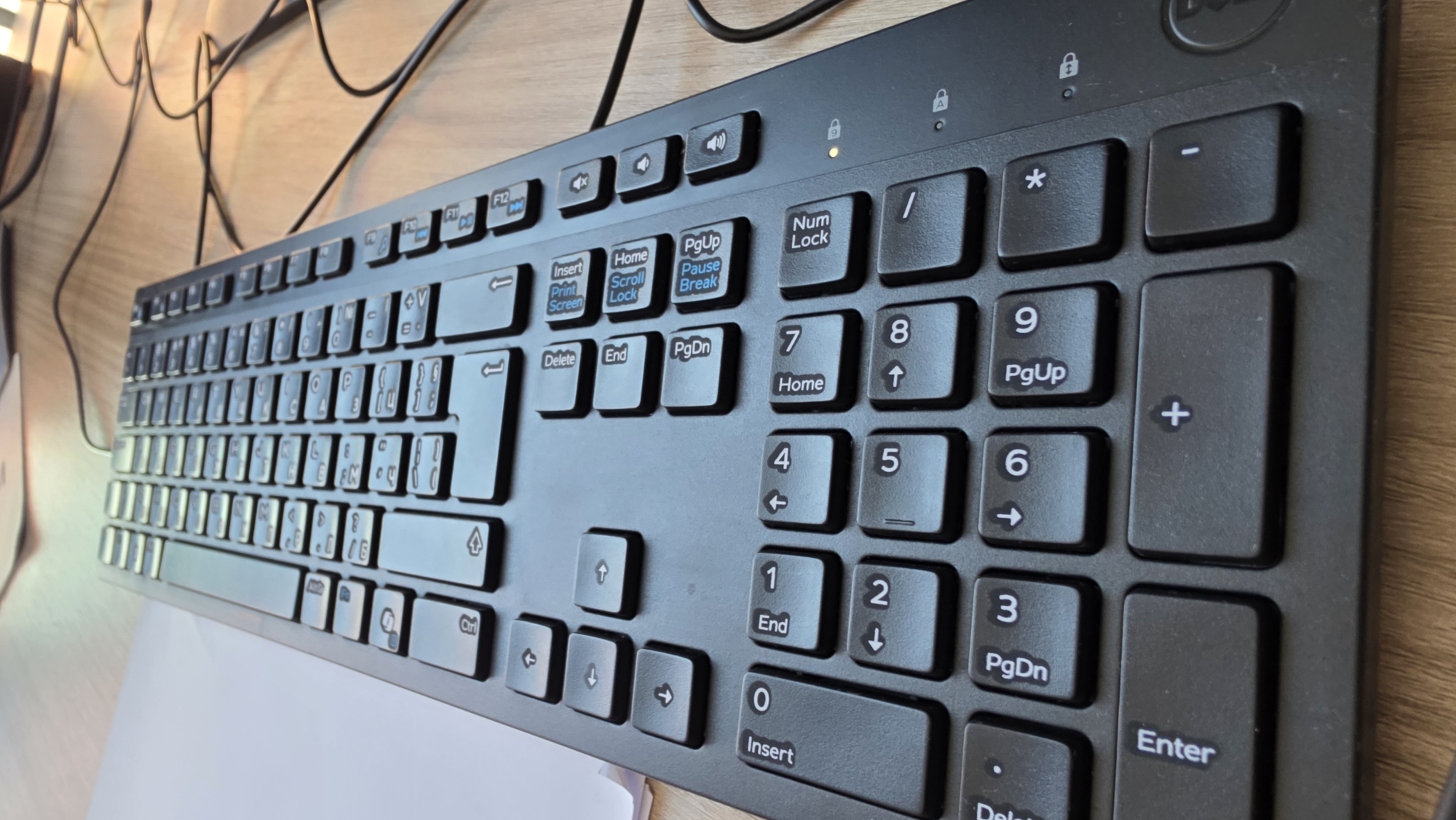 A close-up, overhead shot shows a black computer keyboard on a light brown wooden desk. The keyboard is positioned diagonally from the lower left to the upper right of the frame. The keys are standard QWERTY layout with additional function and navigation keys. Some keys are illuminated with an orange light, indicating activity or status. Several cables are visible trailing from the keyboard to the right side of the frame. The image is taken from a slightly elevated angle, suggesting the camera is held by a person or mounted above the desk. The environment appears to be an office or workspace.