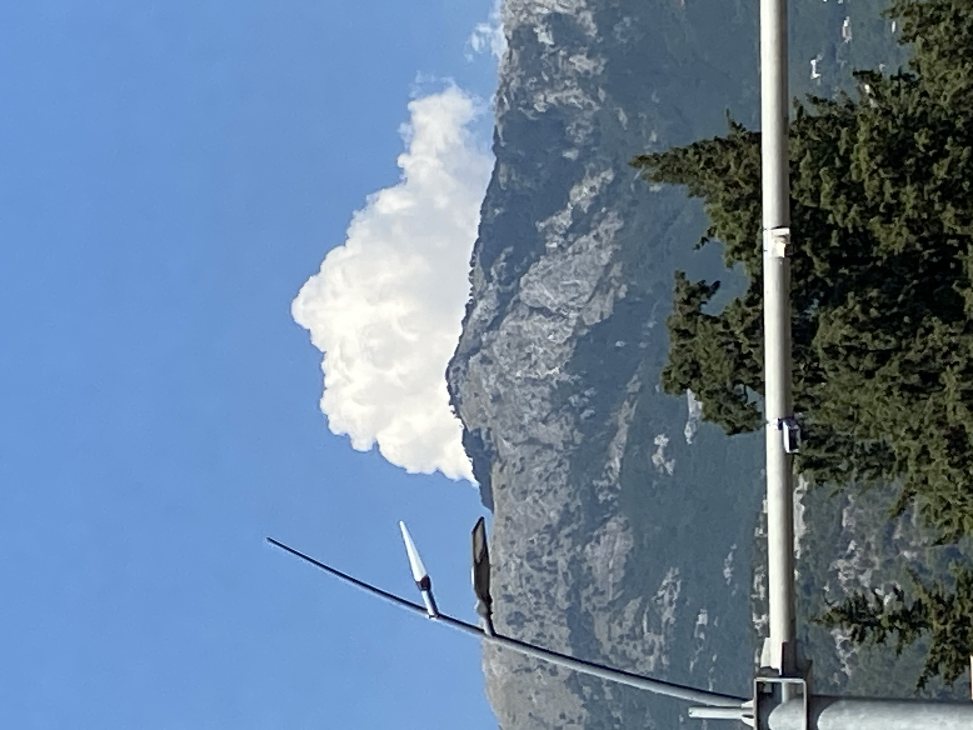 A mountain range, possibly in Budva, Montenegro, is seen under a clear blue sky with a large, fluffy white cloud positioned just above the peak. In the foreground, a street light pole with two lights is visible on the left, and a white horizontal bar extends across the middle of the image. Green trees are scattered across the lower portion of the frame, with a hint of houses in the distant background. The scene appears to be a daytime shot.