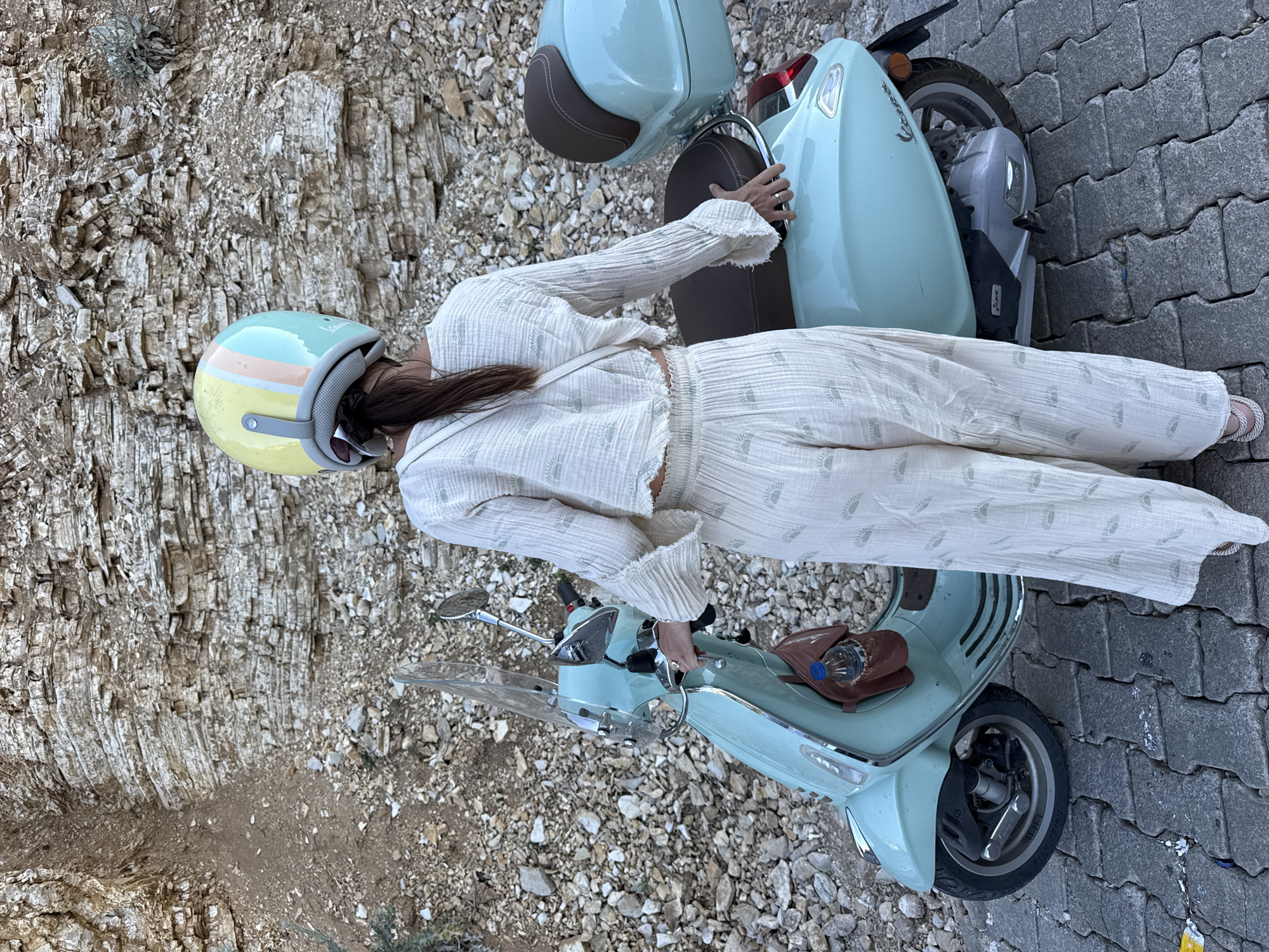 A person wearing a light-colored, loose-fitting outfit adorned with a subtle sun-like pattern and a multi-colored helmet stands next to a light blue scooter. The scooter has a brown seat and a clear windshield. The person's right hand is resting on the scooter's handlebar, and a small brown pouch is visible on the rear of the seat, containing a plastic bottle.
The scene is set outdoors with a rough, rocky terrain to the left and a paved surface composed of dark gray interlocking stones to the right. The rocky area appears to be a cliff face or embankment with layered sedimentary rock. The pavement is clean with a single yellow line visible on the far right.
The lighting suggests it is daytime, with no distinct shadows indicating a specific time. The overall mood seems relaxed and possibly adventurous, given the presence of the scooter and the helmet. The location is identified as Fethiye, Turkey. No visible text is present in the image.