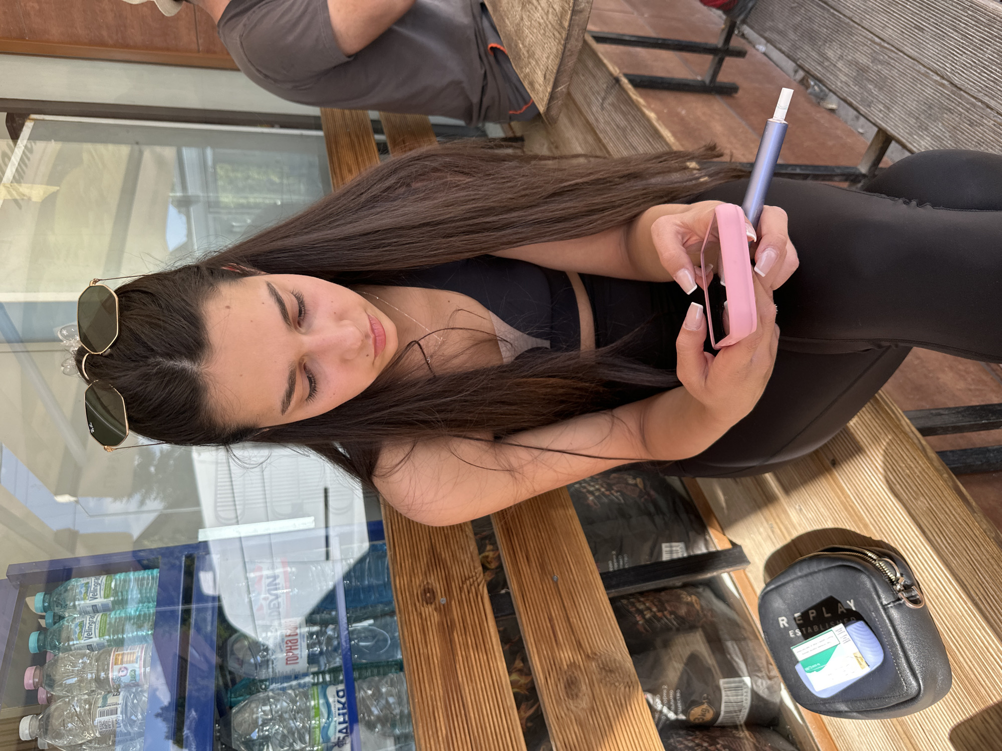 A young woman sits on a wooden bench in Plovdiv, Bulgaria. She holds a pink phone case in her left hand and a blue vaping device in her right. She has long brown hair and wears a black top. Sunglasses rest on her head. A small black purse with the word "REPLAY" is on the bench next to her. In the background, a cooler filled with bottled water is visible. An older man is partially visible at the top of the frame.