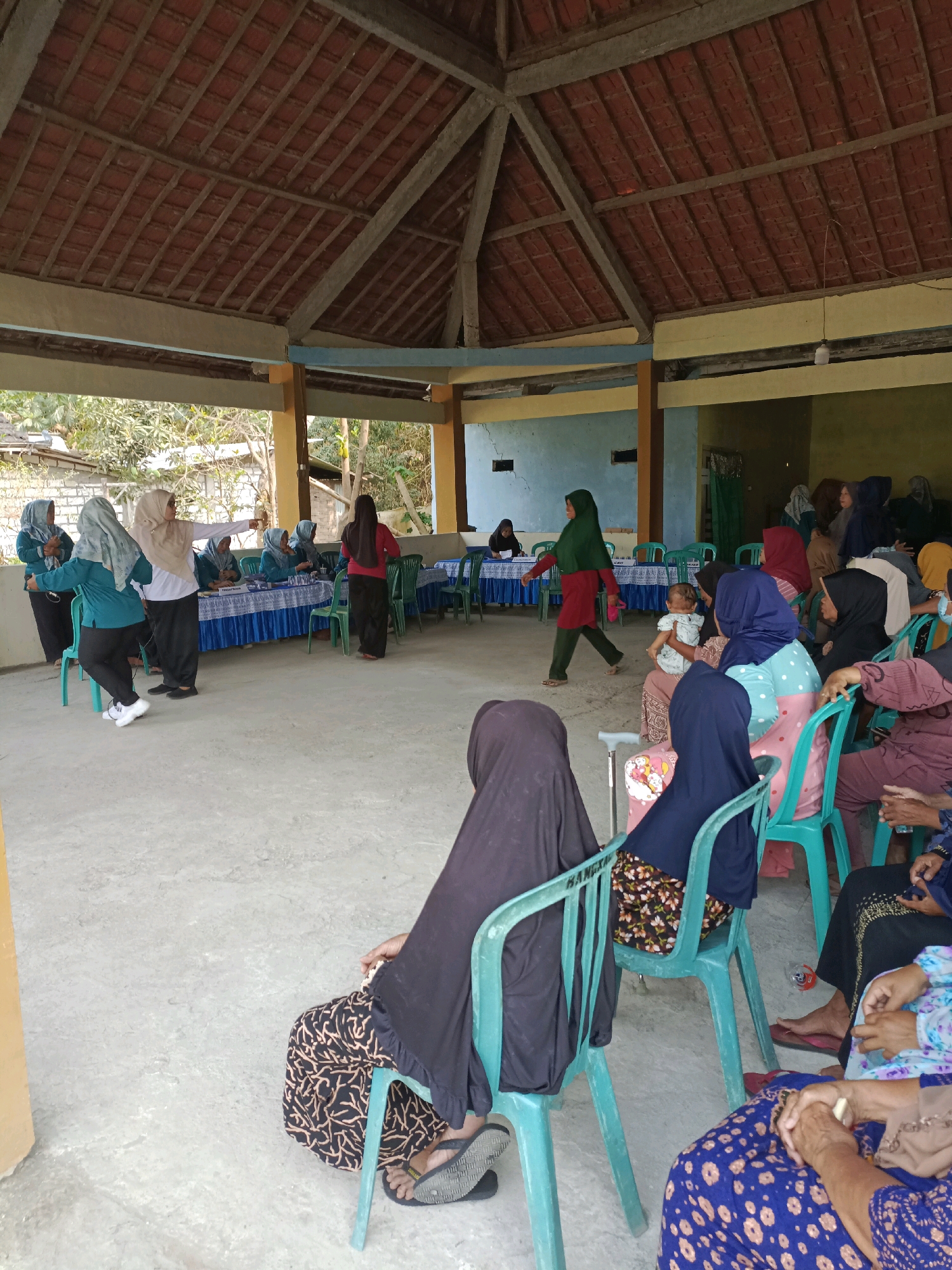 The image depicts an outdoor gathering in Rancang Kencono, Indonesia, under a covered structure with a tiled roof supported by wooden beams. The ground is paved with concrete. In the foreground, several individuals are seated on teal plastic chairs, facing away from the camera. They are wearing traditional Indonesian attire, including hijabs.
In the mid-ground, a group of people are standing or seated around tables covered with blue tablecloths. Some individuals appear to be interacting or engaged in an activity. A woman in a green hijab and red top walks towards the left side of the frame, carrying a pink bag.
Further back, more people are visible, some standing and others seated. The background shows the exterior of buildings and trees, suggesting a semi-rural or communal outdoor space. The overall atmosphere appears to be a local event or meeting.