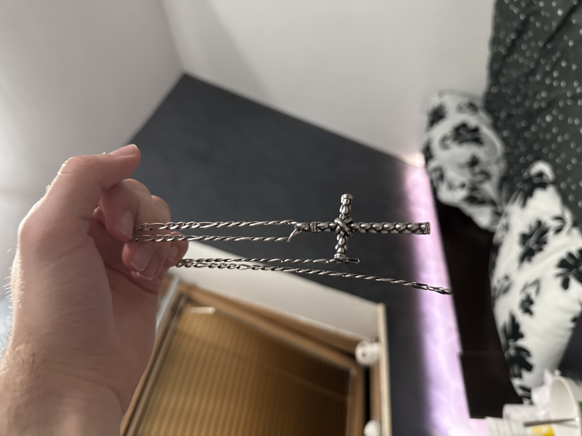 A close-up shot shows a person's hand holding a silver cross necklace. The necklace chain is intricate and appears to be made of twisted metal links. The cross itself is also detailed, with a textured surface. The background is blurred, but it appears to be an indoor setting with a slanted wall and a window with blinds. There is a bed with patterned pillows visible in the lower right portion of the image. The lighting suggests an ambient indoor light source, possibly with a pinkish hue reflecting from below. There are no discernible landmarks or elements that place the image specifically in Blagoevgrad, Bulgaria.