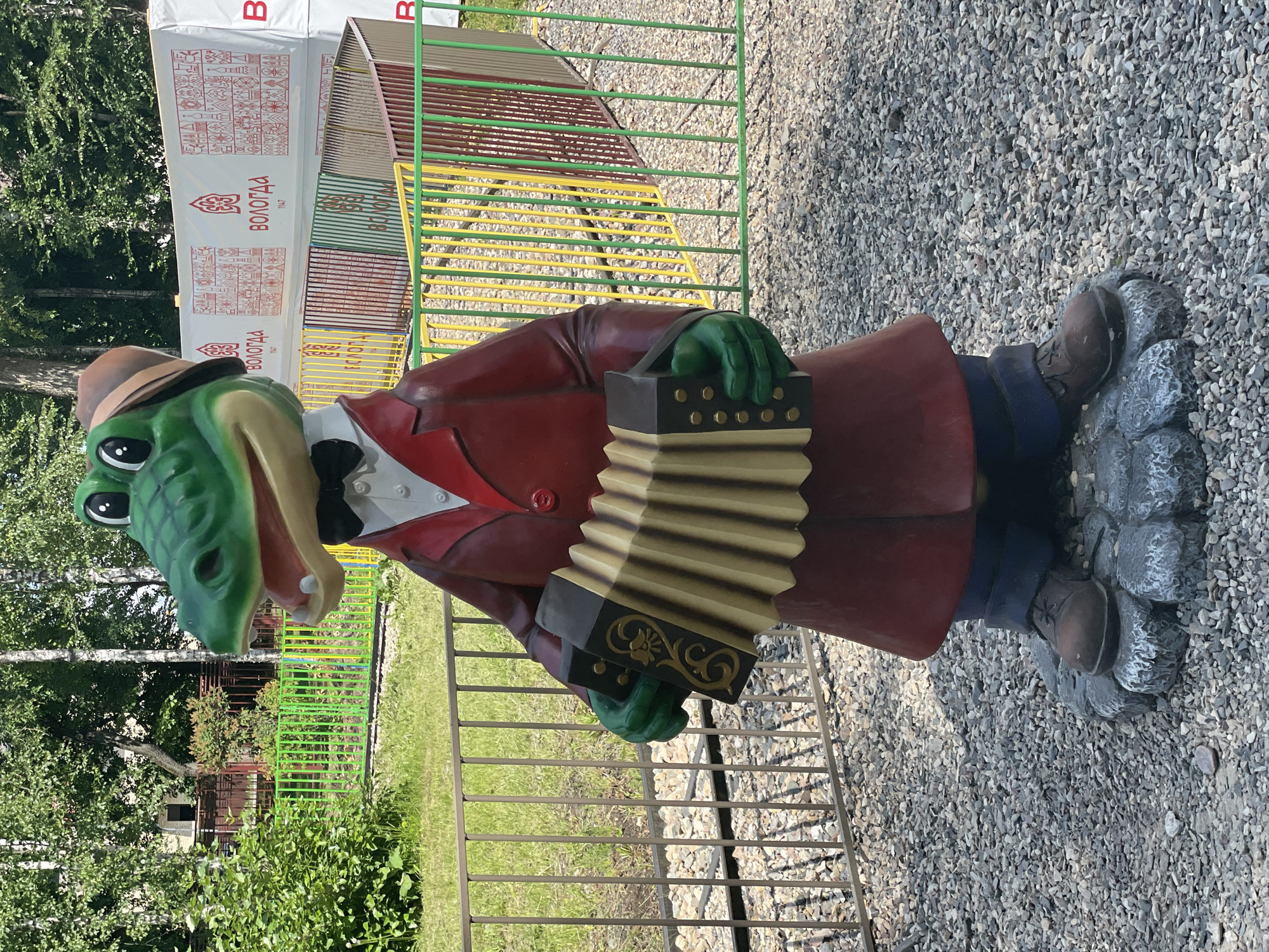 A life-sized statue of a cartoon alligator wearing a red coat, a bow tie, and a hat is depicted playing an accordion. The statue stands on a grey, rock-like base. The alligator is green with a pink mouth and white teeth. The accordion is black and gold with beige bellows. The background includes a metal fence, a lush green grassy area, and trees. In the distance, a white banner with red text and patterns is visible, along with colorful fences and more trees. The ground beneath the statue is covered in small grey stones. The scene appears to be outdoors in a public space, likely an amusement park or recreational area, within the city of Vologda, Russia, as indicated by the text on the banner.