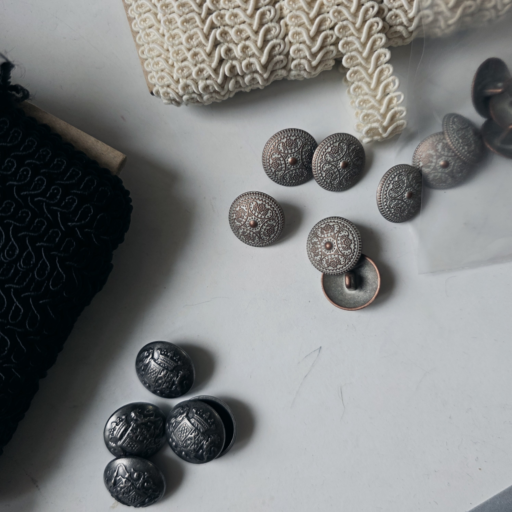 The image presents a top-down, static view of assorted haberdashery items arranged on a light grey, slightly textured surface within an indoor environment in Saint Petersburg, Russia. In the upper left, five oval-shaped, dark metallic buttons are visible, each featuring an intricate, raised crest or emblem design. One button is partially inverted, exposing its hollow rear and shank attachment. Along the top edge, a roll of black textile trim, characterized by a repeating stylized loop pattern, is present, wound around a light brown cardboard core. A second roll of trim, cream-colored and featuring a prominent, complex braided or corded pattern, is positioned vertically along the right side, also wound on a light brown core. Scattered across the mid-right and lower sections are several circular, bronze-toned buttons, each embossed with a detailed floral or geometric design centered around a small dot. One of these buttons is inverted, revealing its concave back and shank. A clear plastic bag partially obscures some of the bronze-toned buttons in the lower right quadrant. The scene is illuminated, casting subtle shadows to the lower right of the objects.