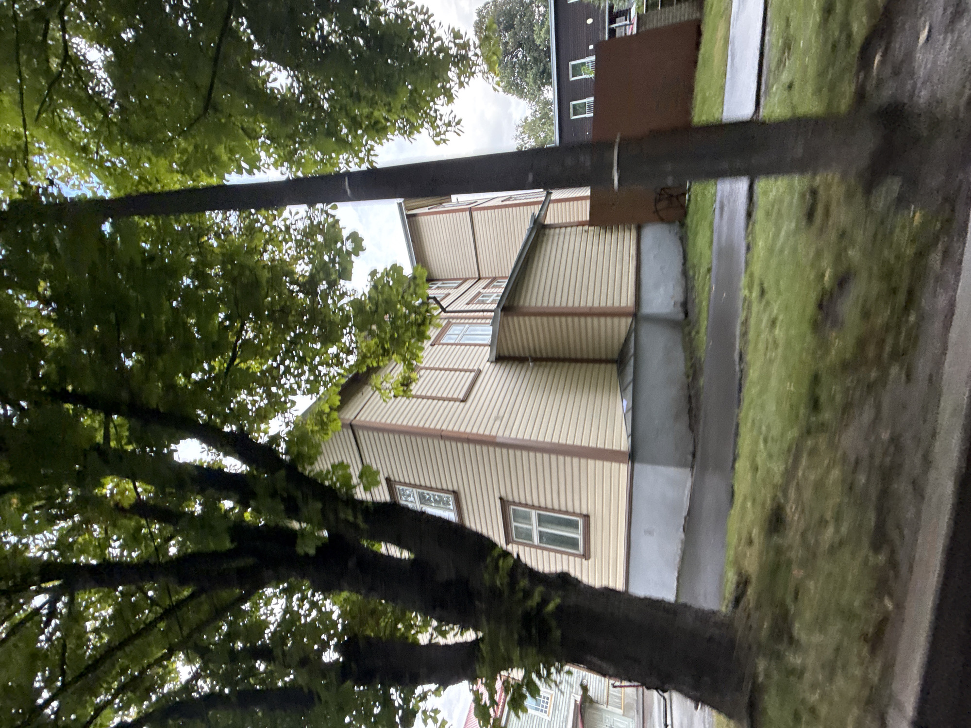 The image captures a side view of a two-story wooden house with cream-colored siding and brown trim, located in Tallinn, Estonia. The house appears to be situated on a street with a sidewalk and a grassy area. A large tree with green leaves partially obscures the front of the house. In the background, another dark-colored building with windows is visible, along with a brown fence or wall. The sky is overcast, suggesting a cloudy day. The perspective of the photo indicates it might have been taken from a moving vehicle.