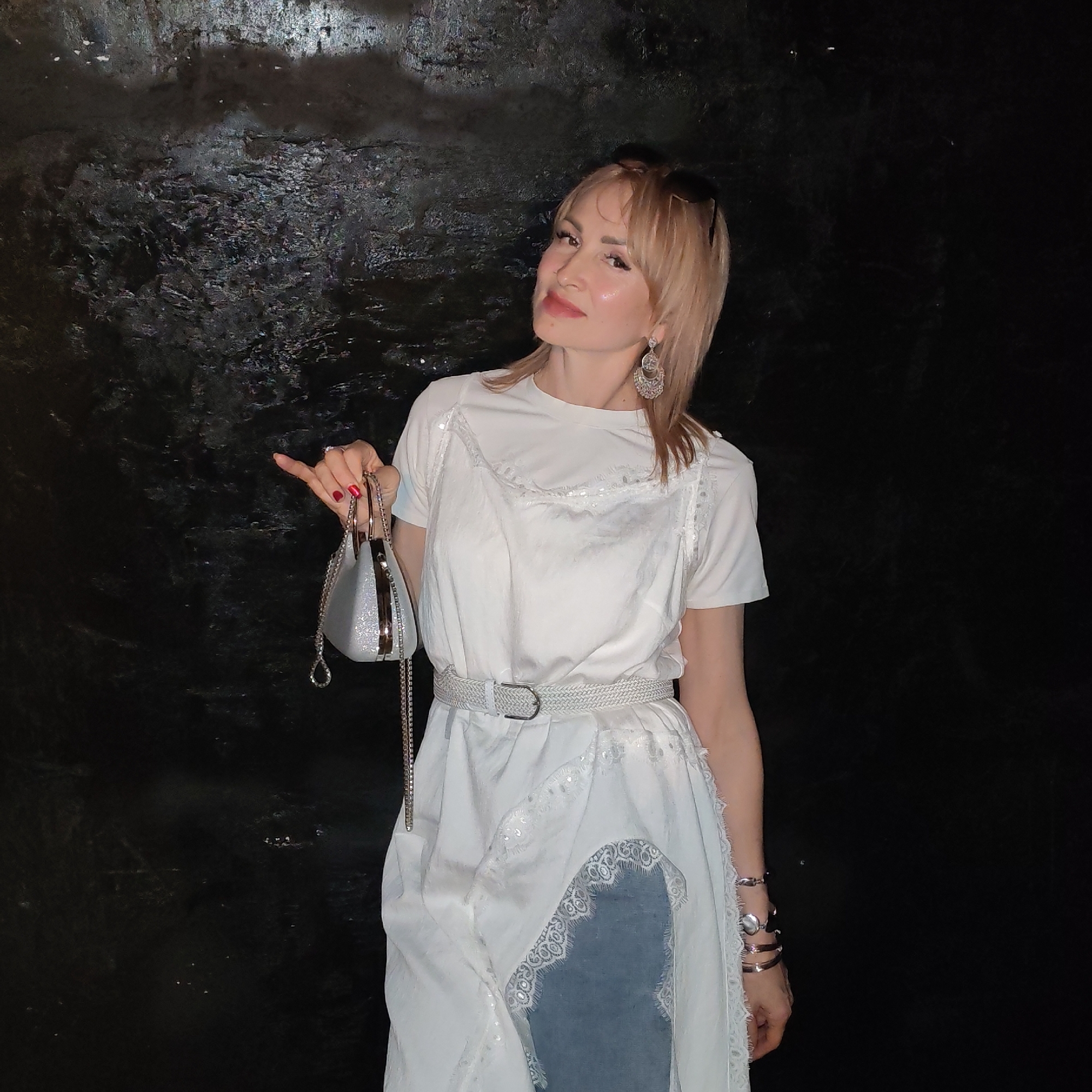A young fair-skinned woman with blonde hair stands against a dark, textured wall. She wears a white t-shirt layered under a white dress with lace trim. A white woven belt accentuates her waist. She holds a small silver purse in her left hand, which has red nail polish. Bracelets adorn her right wrist, and dangling earrings are visible. Sunglasses are perched on top of her head. The location is Chaikovsky, Russia.