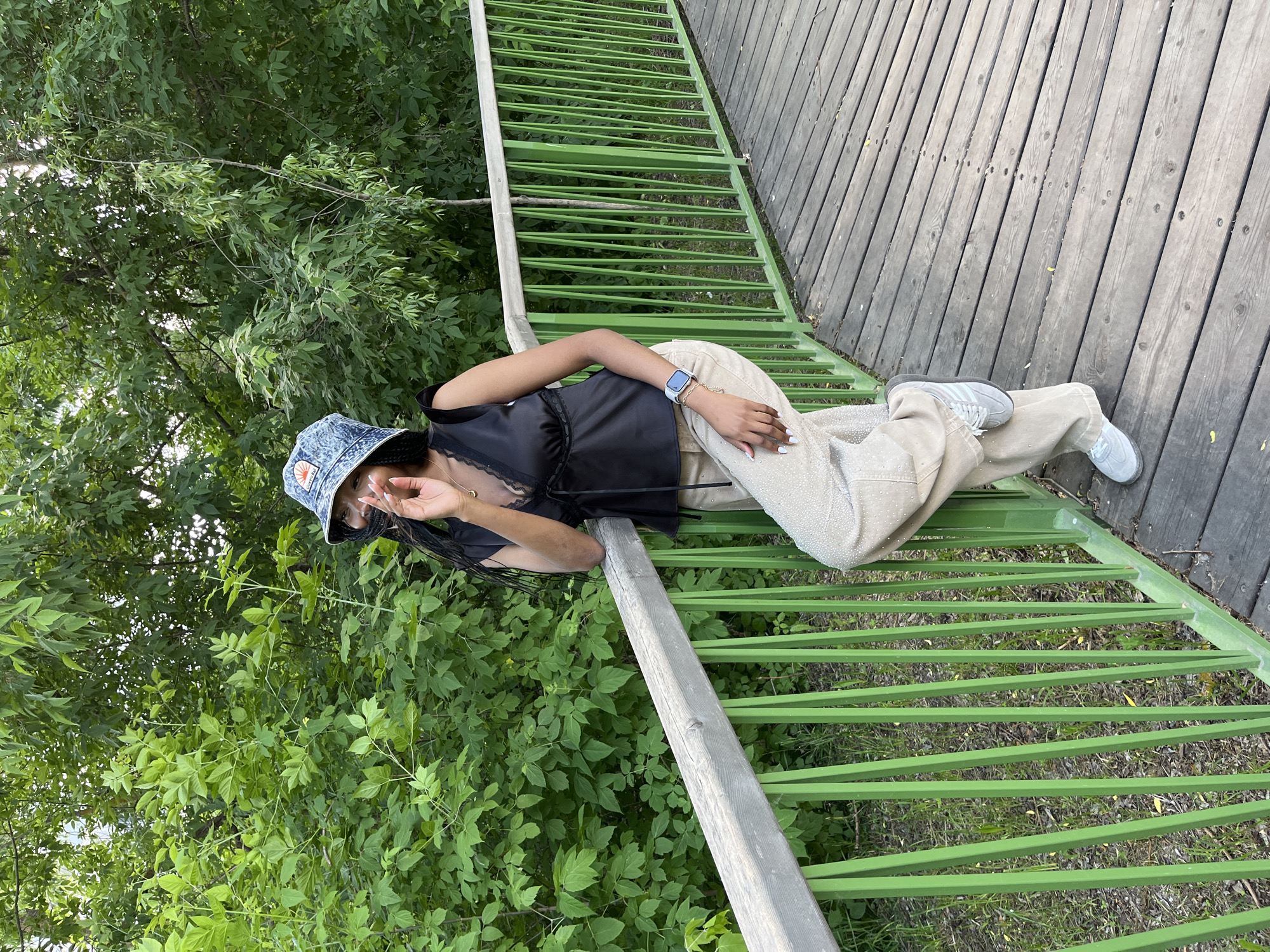 A young Black woman is standing on a wooden boardwalk with a bright green metal railing in a park. She is wearing a denim bucket hat, a black lace-trimmed camisole, and light-colored, loose-fitting pants that appear to be covered in small rhinestones. Her right arm is resting on the railing as she leans against it, and her left hand is raised to her face, with her fingers near her mouth. Her left leg is bent and resting on her right thigh. The boardwalk is made of weathered wooden planks. Behind the railing, lush green foliage forms a dense backdrop, suggesting a natural or park-like setting. The lighting indicates it is daytime, possibly late afternoon, with soft, natural light. The image does not contain any elements that would suggest a specific location in the city of Kazan, Russia.