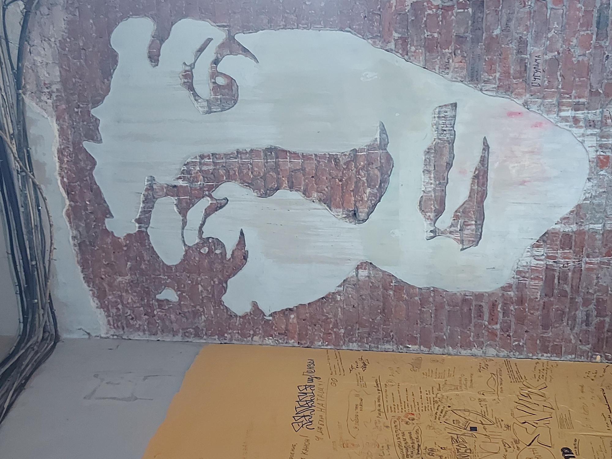 The image shows a portion of a brick wall with a large, irregular, light-colored area that resembles a partially obscured or removed mural. The brick wall is aged, with some areas of exposed brick and others covered by plaster or paint. There are black wires running vertically on the left side of the wall. A large sheet of tan paper with handwritten text and doodles lies on the floor in front of the wall. The text appears to be in Russian. Based on the context of the text on the brick wall reading "В УРУЧЬЯ" the location is likely to be near the city of Saint Petersburg in Russia, specifically in or around a place called Uruchye. The scene appears to be inside a building under construction or renovation, with visible electrical wiring and an unfinished wall surface. The overall impression is of a space that is in a state of transition or decay.
