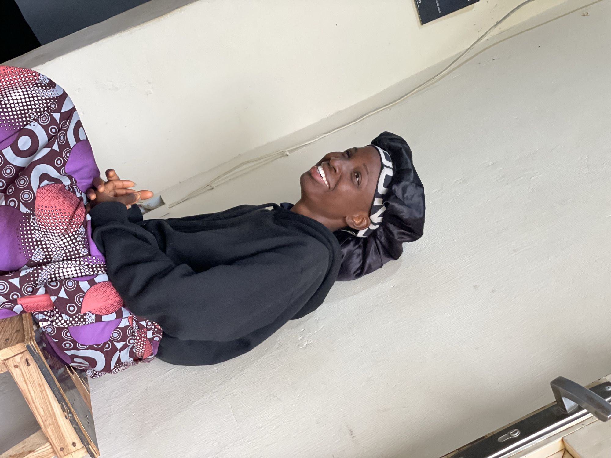 A young Black female is sitting on a wooden stool against a light-colored wall. She is wearing a black hoodie and a black satin bonnet with a white and black geometric pattern around the brim. She is also wearing a brightly colored patterned skirt with shades of purple, pink, and white. Her hands are clasped in her lap, and she is looking to her left with a wide smile. The background wall appears to be textured, and a light-colored electrical wire runs down the wall. To the right, a door handle and a portion of a door frame are visible. The overall lighting suggests an indoor or shaded outdoor environment. The image is set in Durumi, Nigeria.