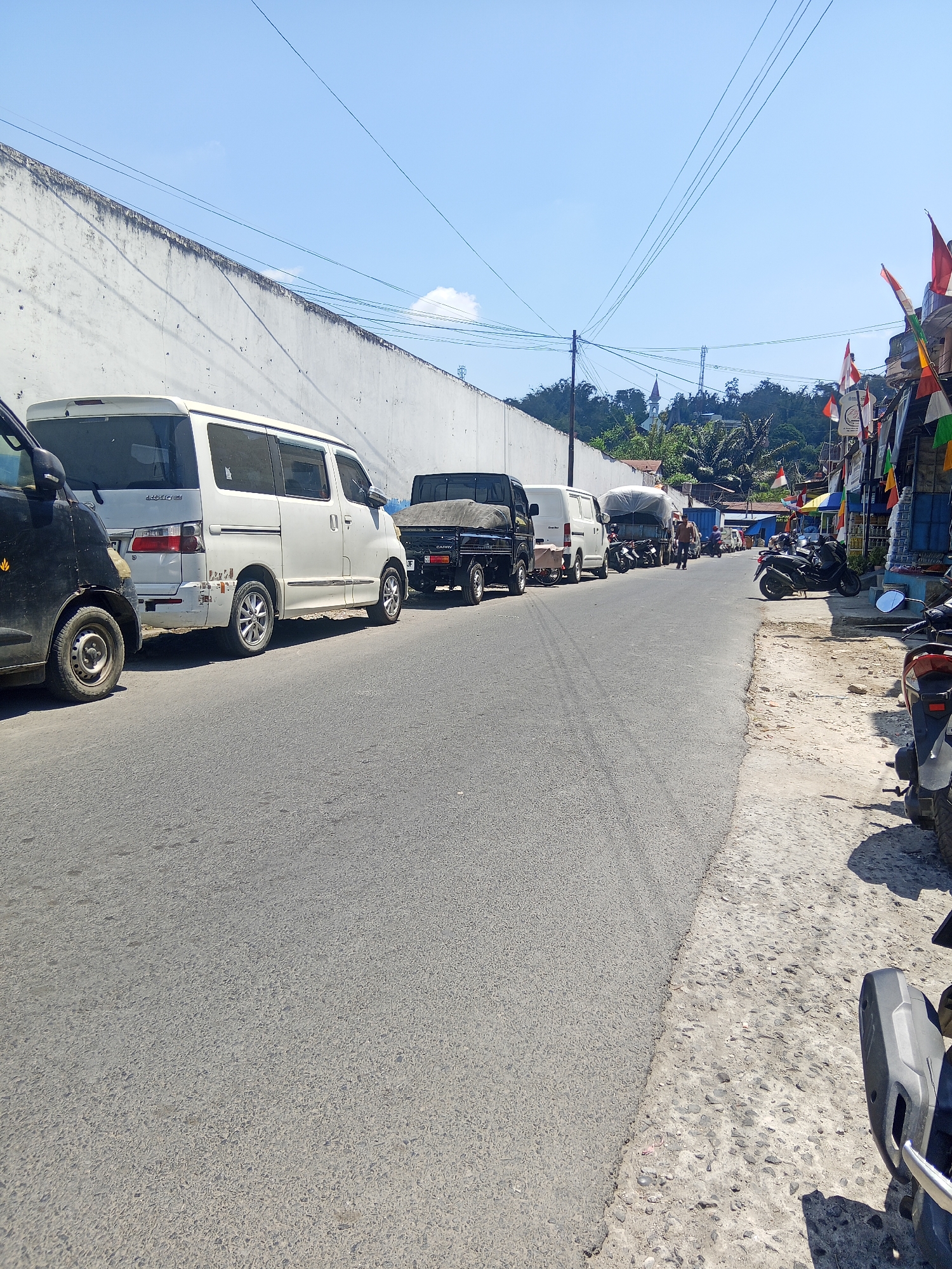 This is a street scene in Makale, Indonesia, on a bright, sunny day. The street is paved with asphalt and bordered by a rough, unpaved shoulder. On the left side of the street, a white van and a black pickup truck are parked, partially obstructing a high, white, textured wall. Further down the street, several other vehicles are parked, including motorcycles, another white van, and a covered truck. On the right side of the street, more motorcycles are parked, and there appears to be a shop with various colorful flags and awnings. In the background, a hillside is visible with trees, buildings, and what looks like a church steeple and a telecommunications tower. A few people are seen walking on the street. Overhead, power lines crisscross the sky.
