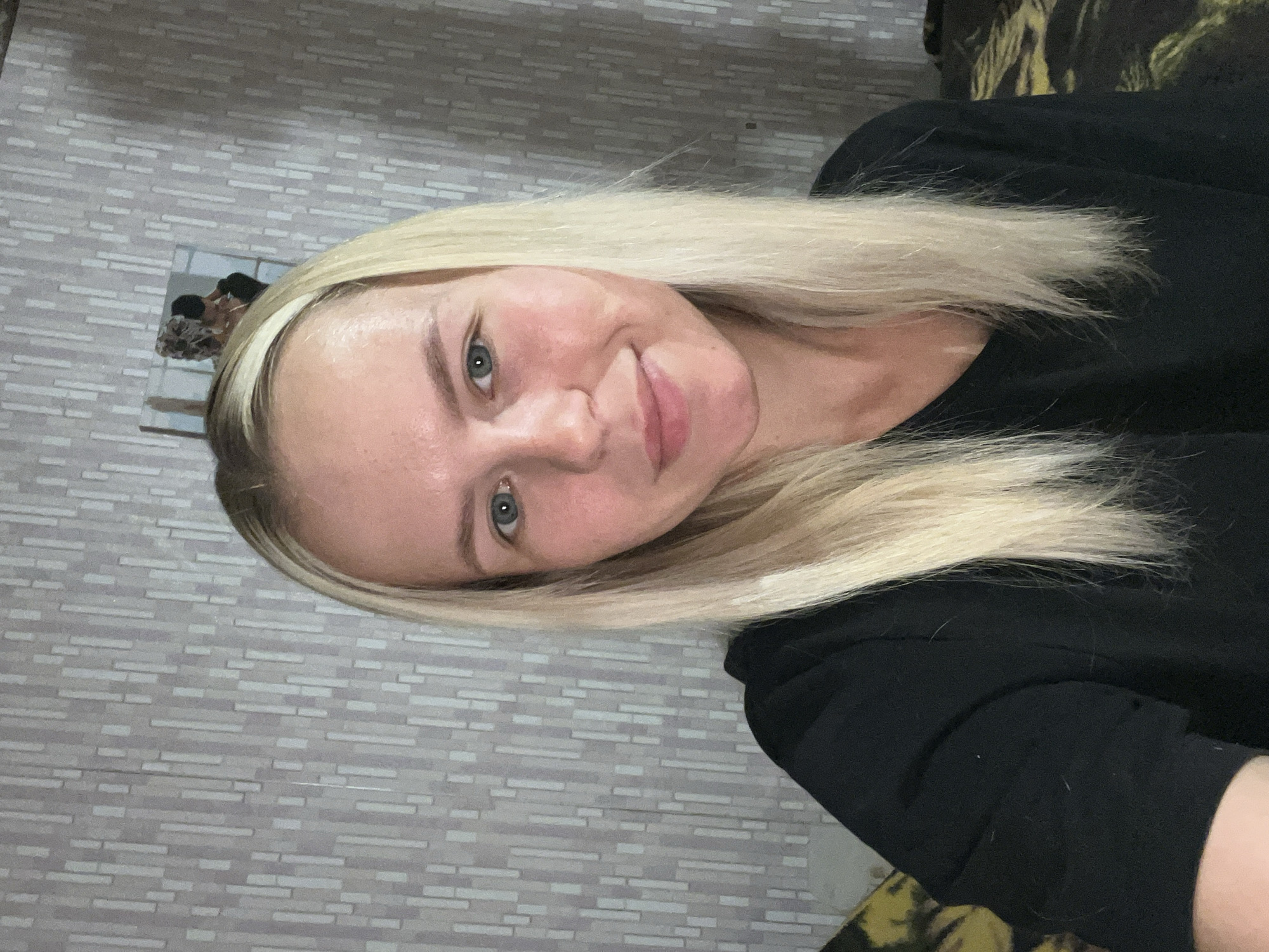 A young woman with blonde hair and blue eyes is pictured in a close-up, self-taken photograph. She is wearing a black t-shirt and has a light complexion. The background consists of a wall with a vertical striped pattern and a framed picture hanging on it. A portion of a bed with a patterned cover is visible in the lower left of the frame. The scene appears to be indoors.