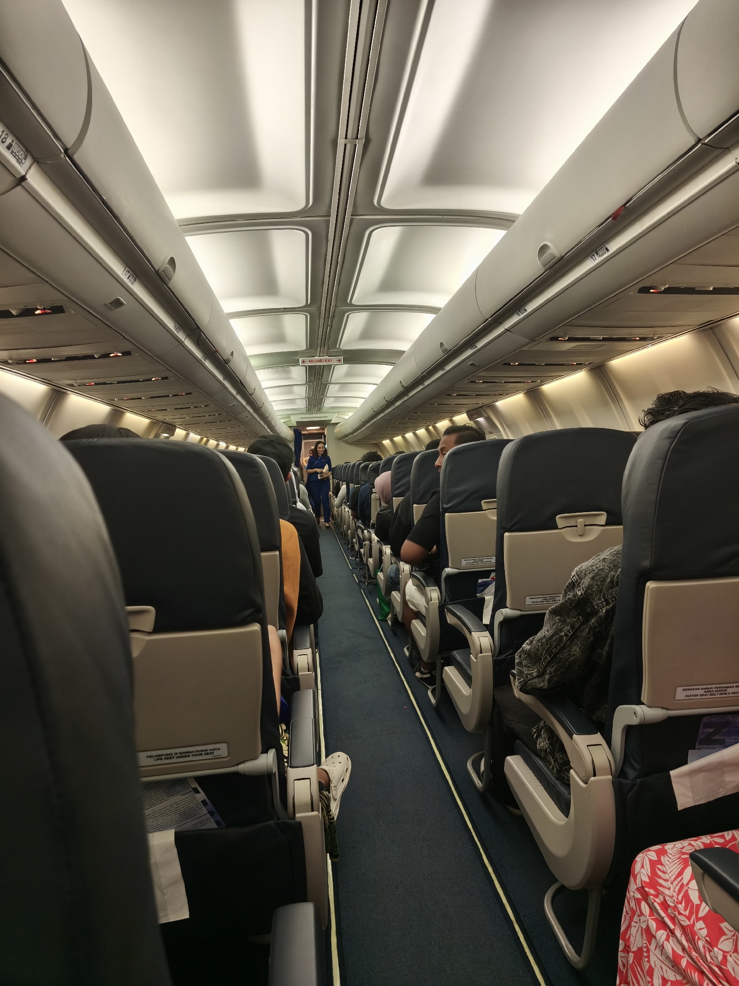 The image depicts the interior of an airplane cabin, viewed from a low angle, looking forward down the aisle. The cabin is lit by overhead lights. Rows of dark-colored airline seats with beige headrests are visible on both sides of the aisle, some occupied by passengers whose heads and shoulders are visible. The aisle itself is carpeted in dark blue with a yellow stripe running down its center. Further down the aisle, a flight attendant in a blue uniform is standing and facing the camera, seemingly addressing passengers. Signs for "KELUAR/EXIT" are visible on the right-hand side wall. The overhead compartments are open in some sections. The scene is devoid of any specific Kuta, Indonesia landmarks or features; the setting is a generic airplane interior.