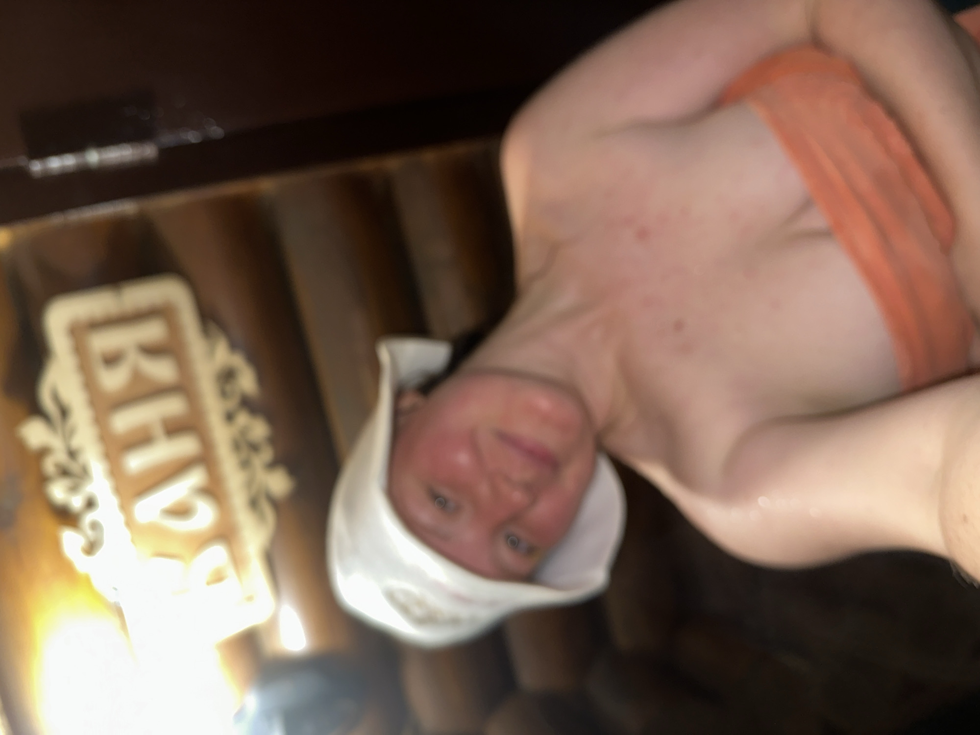 A person in a white sauna hat and orange towel is in a sauna with wooden walls and a sign that says "Баня" in Russian. The location is likely in Nizhny Novgorod, Russia, given the sauna's Russian name and architecture. The person appears to be sweating and flushed, likely due to the high temperature of the sauna.