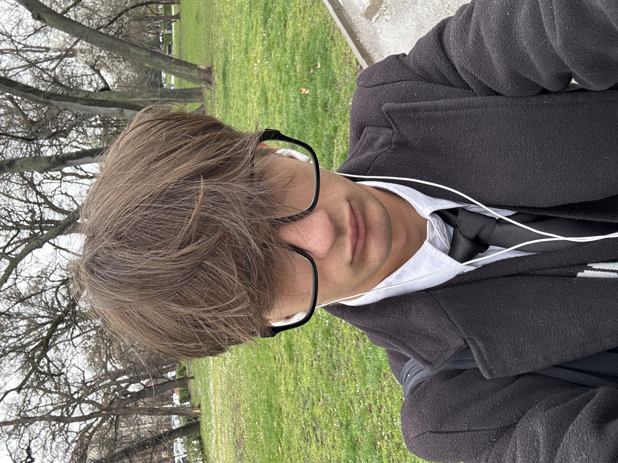 A young man with brown hair and glasses is standing in a park, possibly in Sofia, Bulgaria, given the presence of trees with bare branches common in colder climates. He is wearing a black coat over a white shirt and a black tie. He has earbuds in. The background shows green grass and trees. The image appears to be taken outdoors during the daytime.