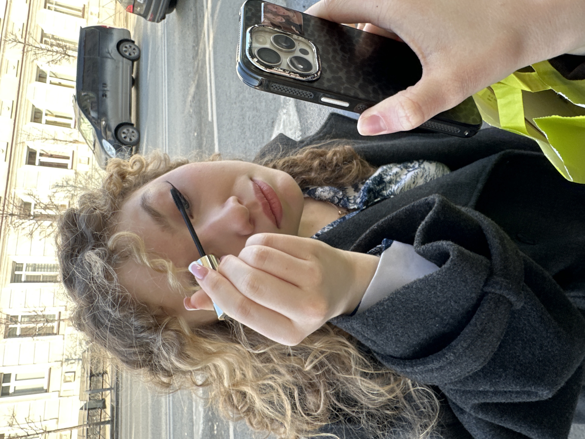 A female individual is depicted in a close-up, outdoors, applying mascara to her right eye. She has light brown, curly hair and is wearing a dark grey overcoat over a white collar and a blue and white patterned garment. Her left hand holds a black smartphone, likely an iPhone 12/13/14/15 Pro Max model, featuring a multi-lens camera array and a dark patterned case with a small rectangular photo affixed to its back. The phone's screen appears to be used as a mirror. Her right hand holds a mascara wand, with the tube visible, exhibiting a metallic gold base and an iridescent light blue/silver top. Her fingernails have a French tip manicure. A neon green shopping bag is partially visible in the lower right, held by the same hand as the phone.
The background shows an urban street scene. Behind the individual, an asphalt road with white lane markings is visible, upon which a dark grey Hyundai Starex/H-1 minivan is parked or driving. Another vehicle, a dark sedan, is partially visible behind the minivan. Across the street, a light-colored, multi-story building with classical architecture, featuring multiple rectangular windows with white frames, stands. Bare deciduous trees are situated along the street between the building and the road. The scene is illuminated by natural daylight.
The location is a historical street in the central part of Kazan, Russia, identifiable by the architectural style of the background building and the urban infrastructure.